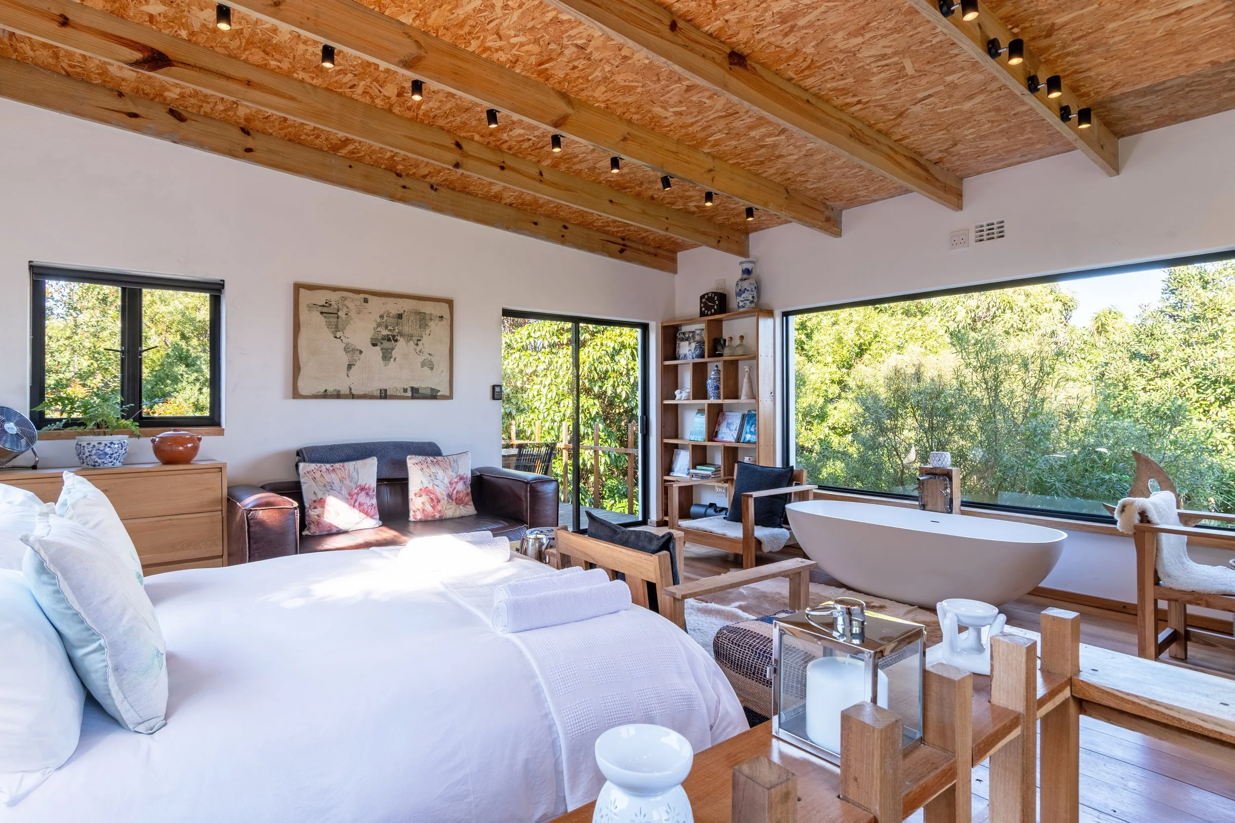 Bright bedroom with a large window view of green trees, a white freestanding bathtub near the window, a bed with white linens, and wooden furniture and ceiling beams, decorated with a world map and various ceramic vases.