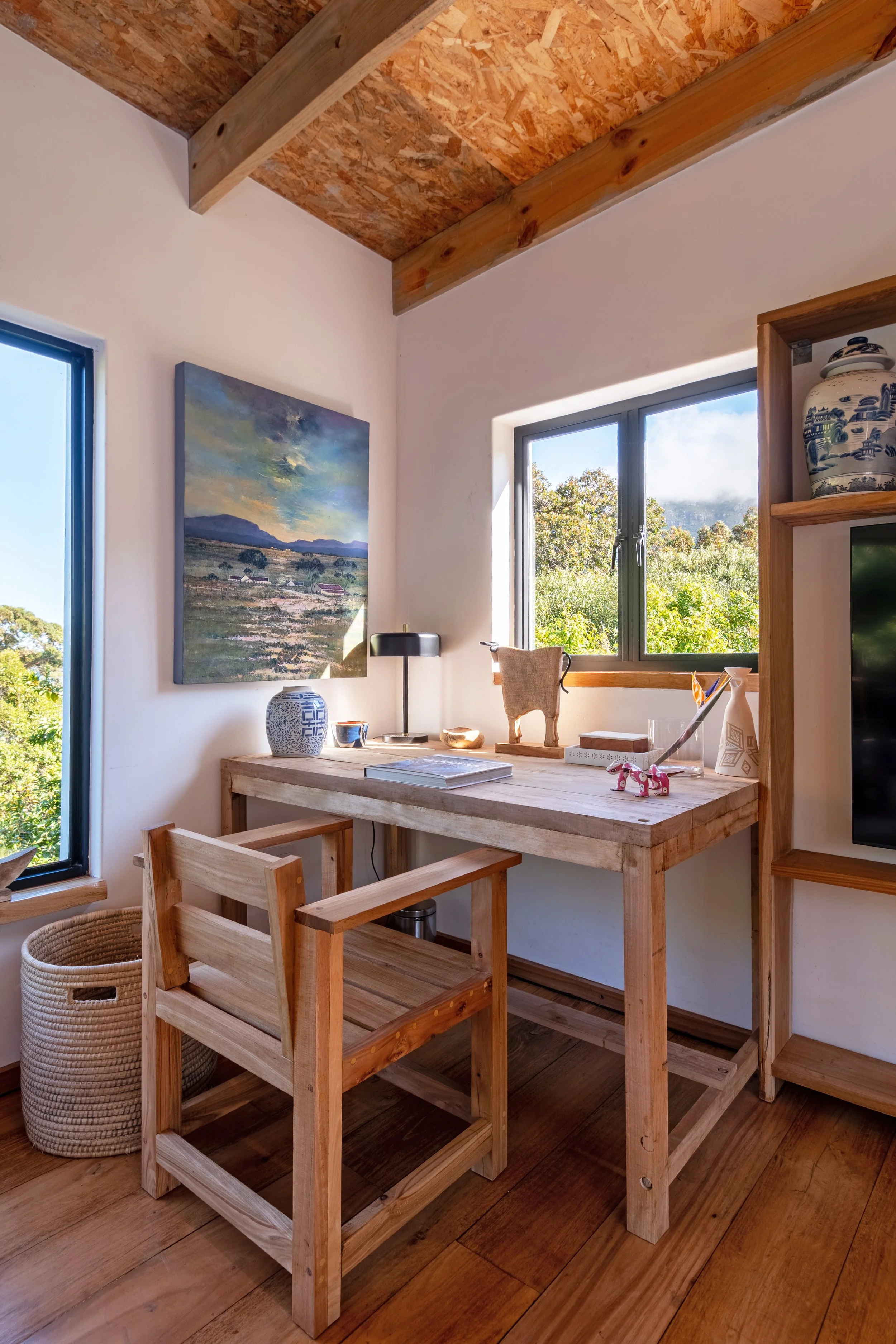 A rustic corner of a room with a wooden desk, matching chair, a window showing greenery outside, and decorative items like ceramics, sculptures, and artwork.