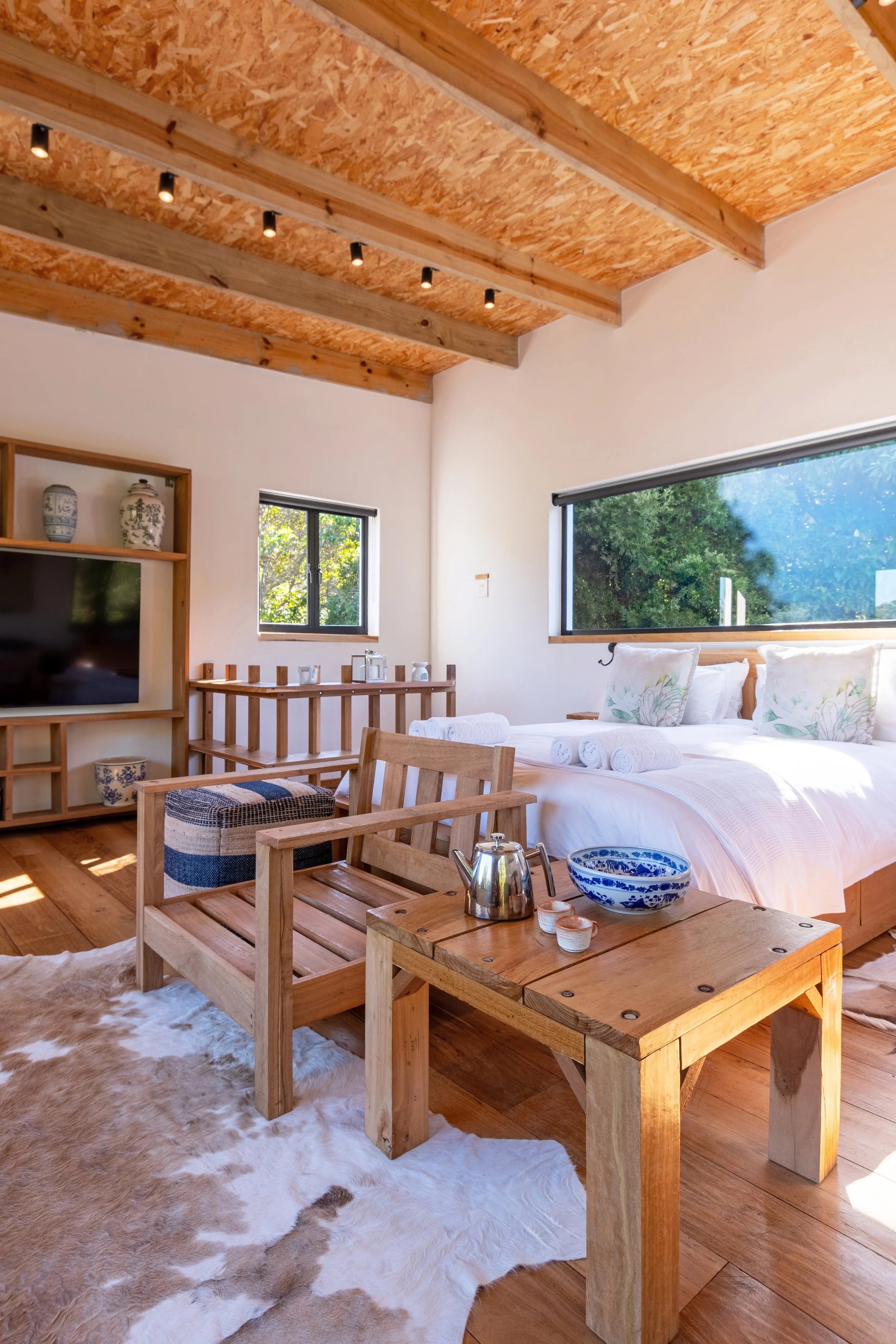 A cozy bedroom with a wooden ceiling, large windows, a bed with white linens and pillows, a small wooden side table with a teapot and bowls, a wooden bench, and a cowhide rug.