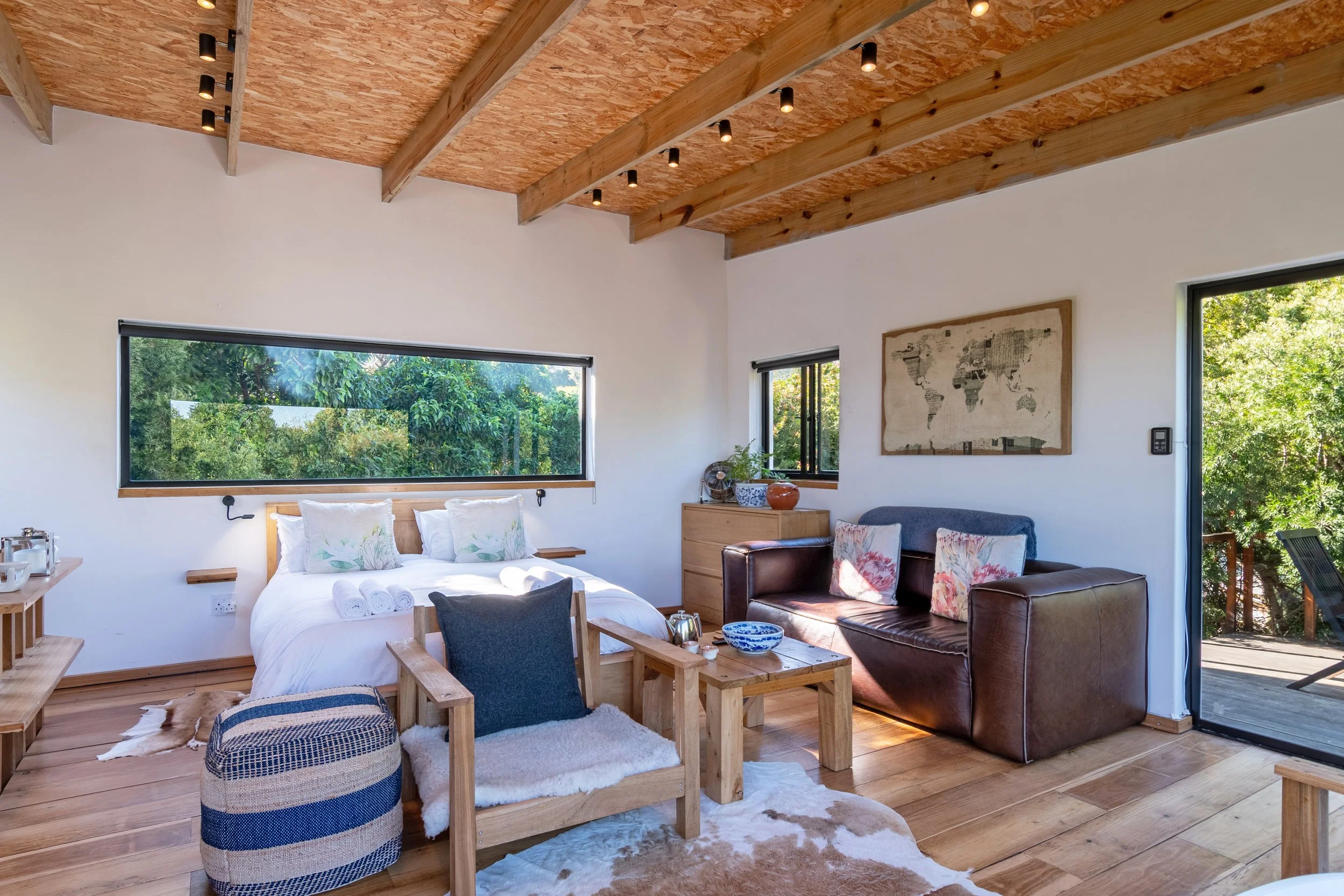 A cozy bedroom with wooden beams and white walls, featuring a double bed with white bedding, a wooden armchair with a black cushion, a small wooden coffee table with a bowl and candles, a leather sofa with floral pillows, and a wooden dresser, with l