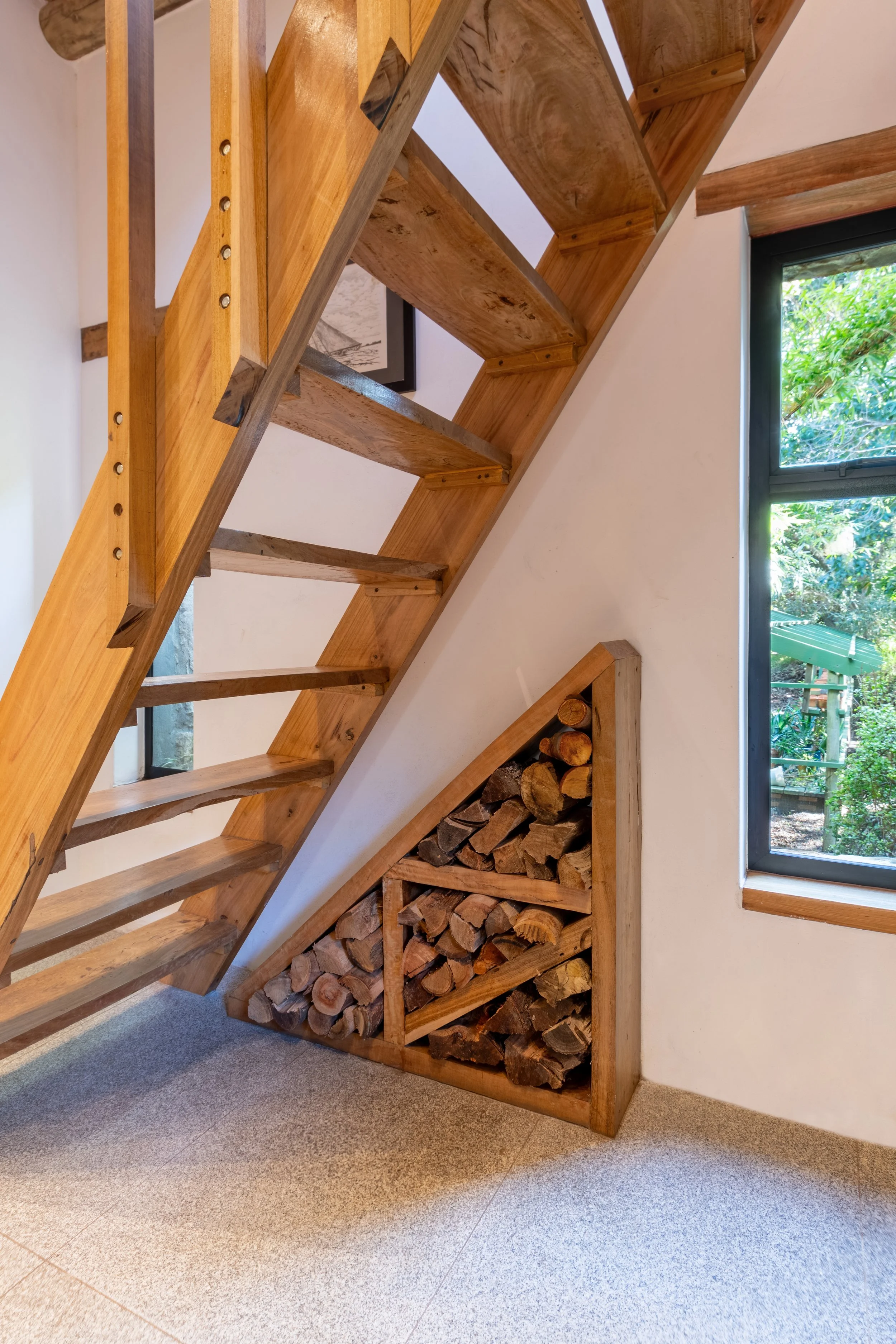 Wooden staircase with a built-in firewood storage area underneath, next to a window showing a green outdoor scene.