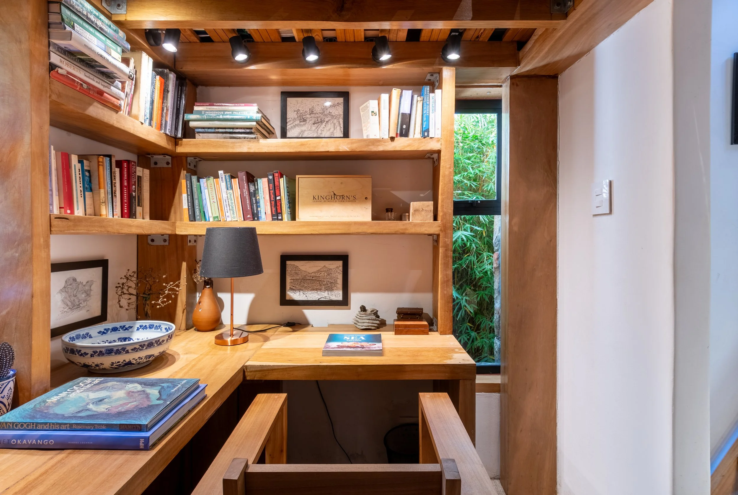A cozy home office nook with wooden shelves filled with books, a small desk with a lamp, and framed artwork, next to a window with a view of green foliage.