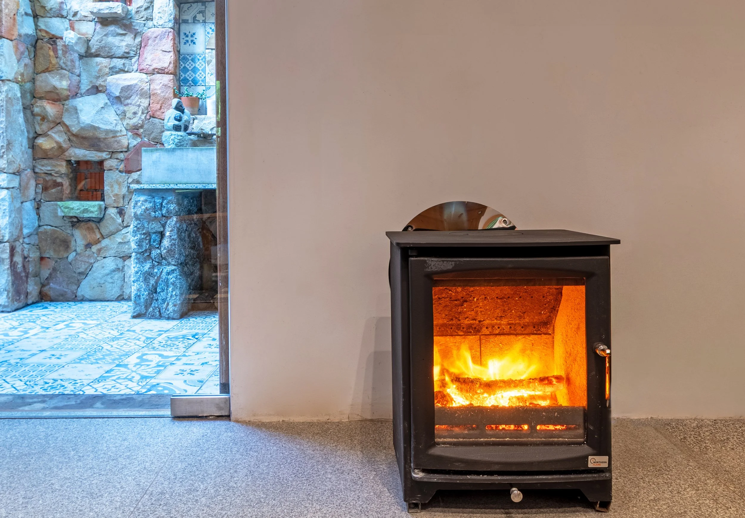 A small black stove with glass door showing a fire inside, placed against a plain wall. To the left, there is a glass door leading outside, revealing a stone wall and patterned tiled floor.