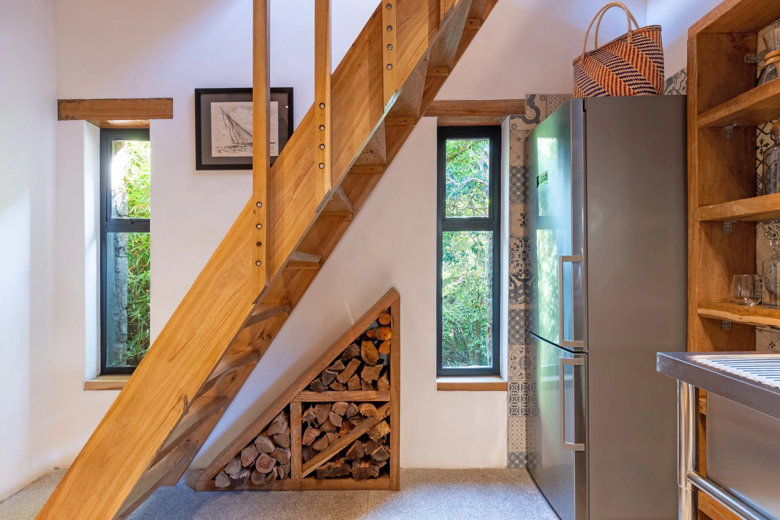 A cozy kitchen with wooden accents, a silver refrigerator, open shelving, two small windows, and a staircase with firewood stored underneath.