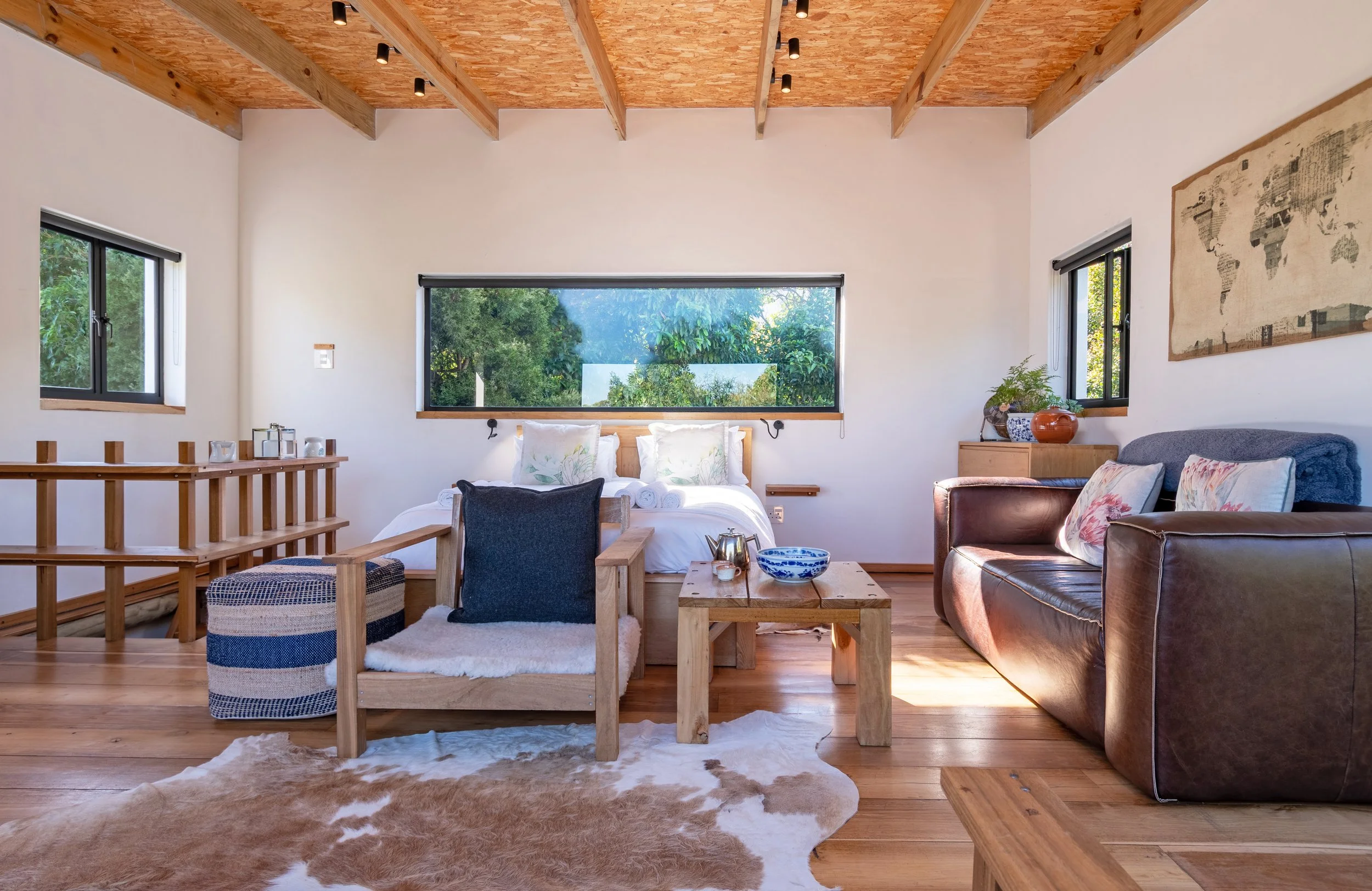 Living room with wooden ceiling beams, large windows showing greenery, a bed with pillows, a brown leather sofa with decorative pillows, a wooden armchair with cushion, a wooden coffee table with a teapot and bowl, and a cowhide rug.