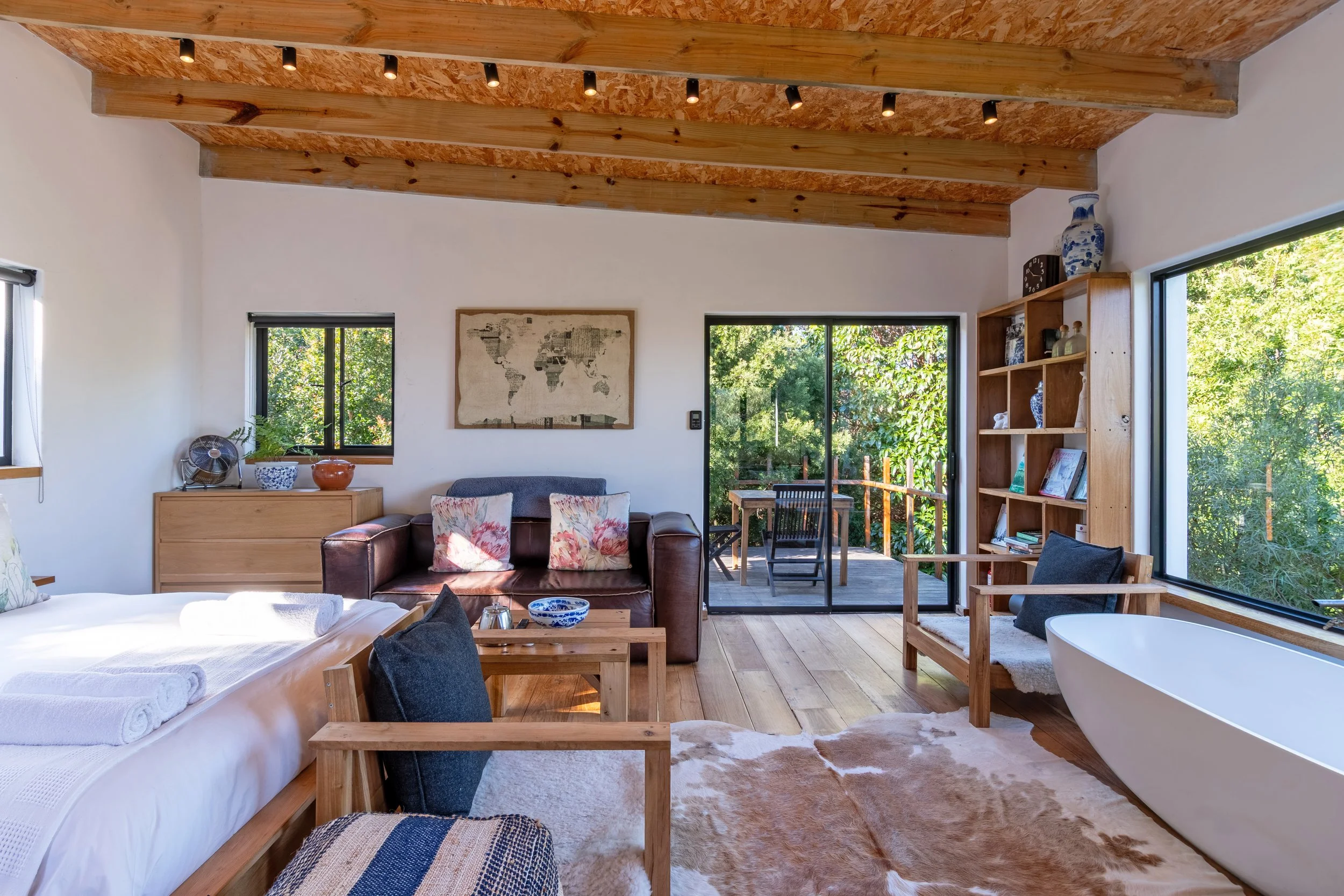 Interior of a cozy living space with a wooden ceiling, large windows, a bed, a leather sofa with floral pillows, a wooden bookcase, and a freestanding bathtub next to the window, with a patio and greenery outside.