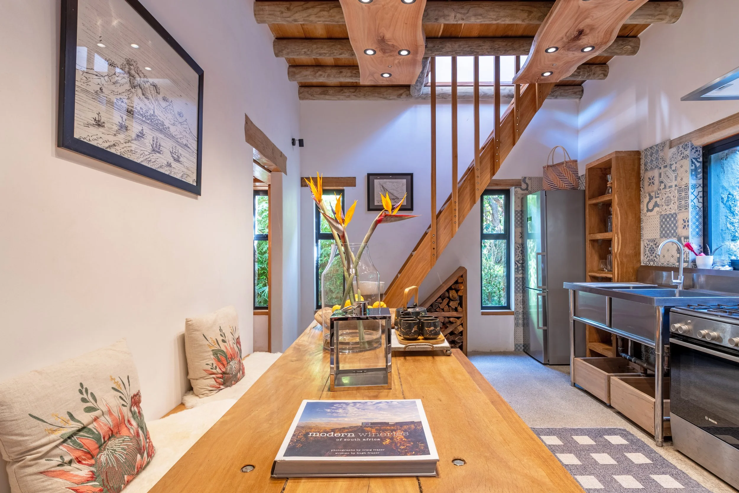 Modern kitchen with wooden accents, a staircase, stainless steel appliances, and a dining area with cushions and a table decorated with a flower arrangement.