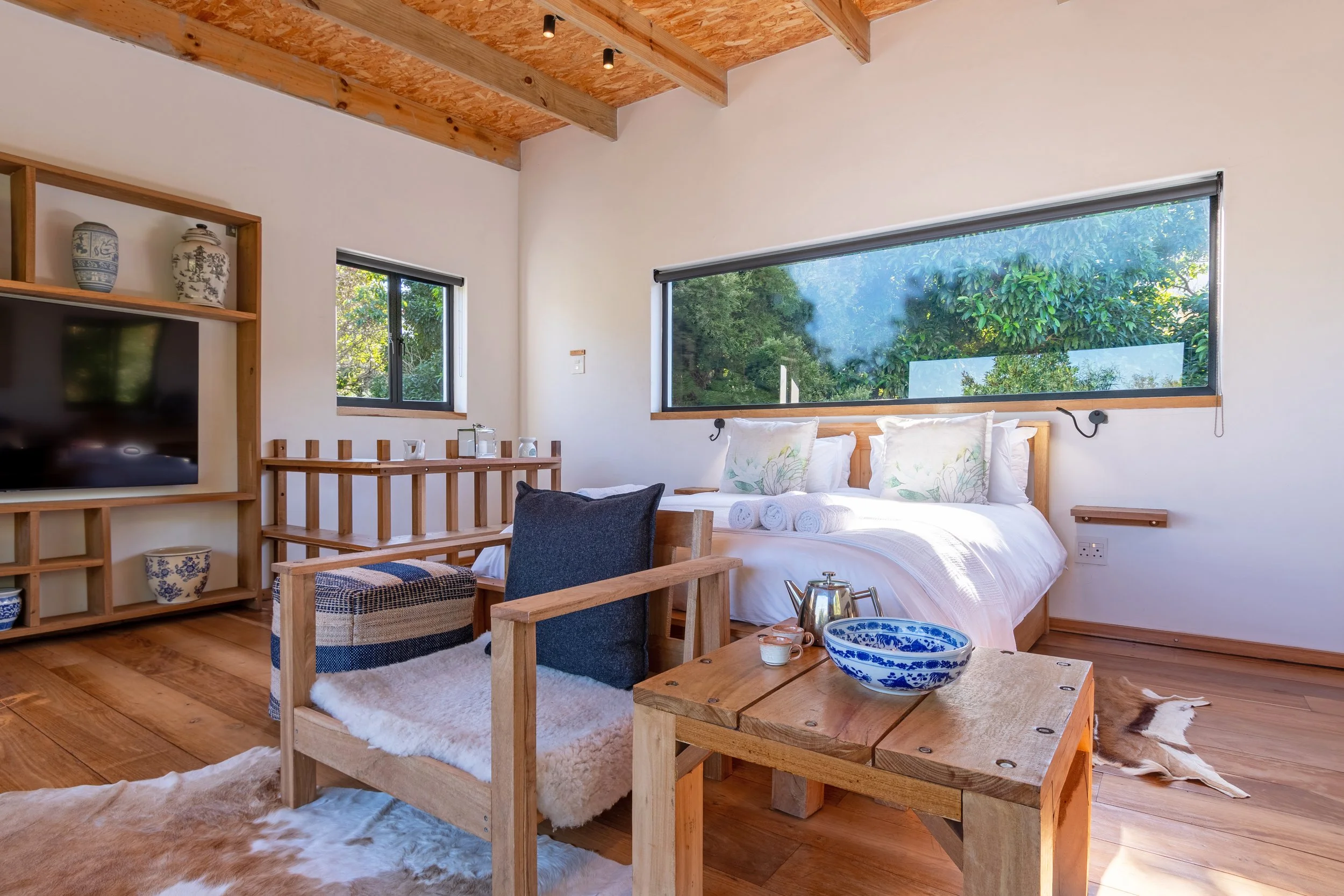 A cozy bedroom with a wooden ceiling, white walls, and large windows showing greenery. It features a bed with white bedding, decorative pillows, a wooden headboard, and rolled towels. There is a armchair with a pillow, a wooden coffee table with a bo