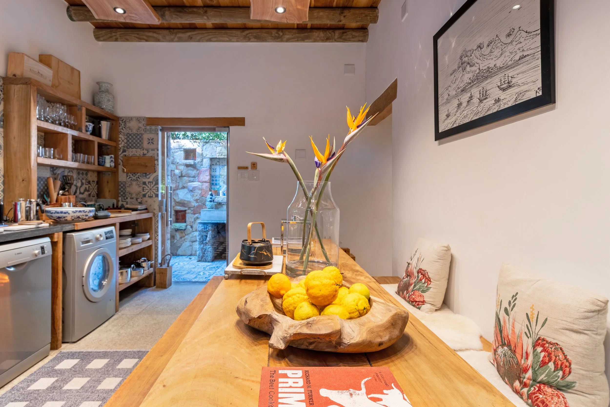 Wooden dining table with a glass vase holding tropical flowers, a wooden bowl filled with lemons and limes, with a kitchen area in the background and a door leading outside.