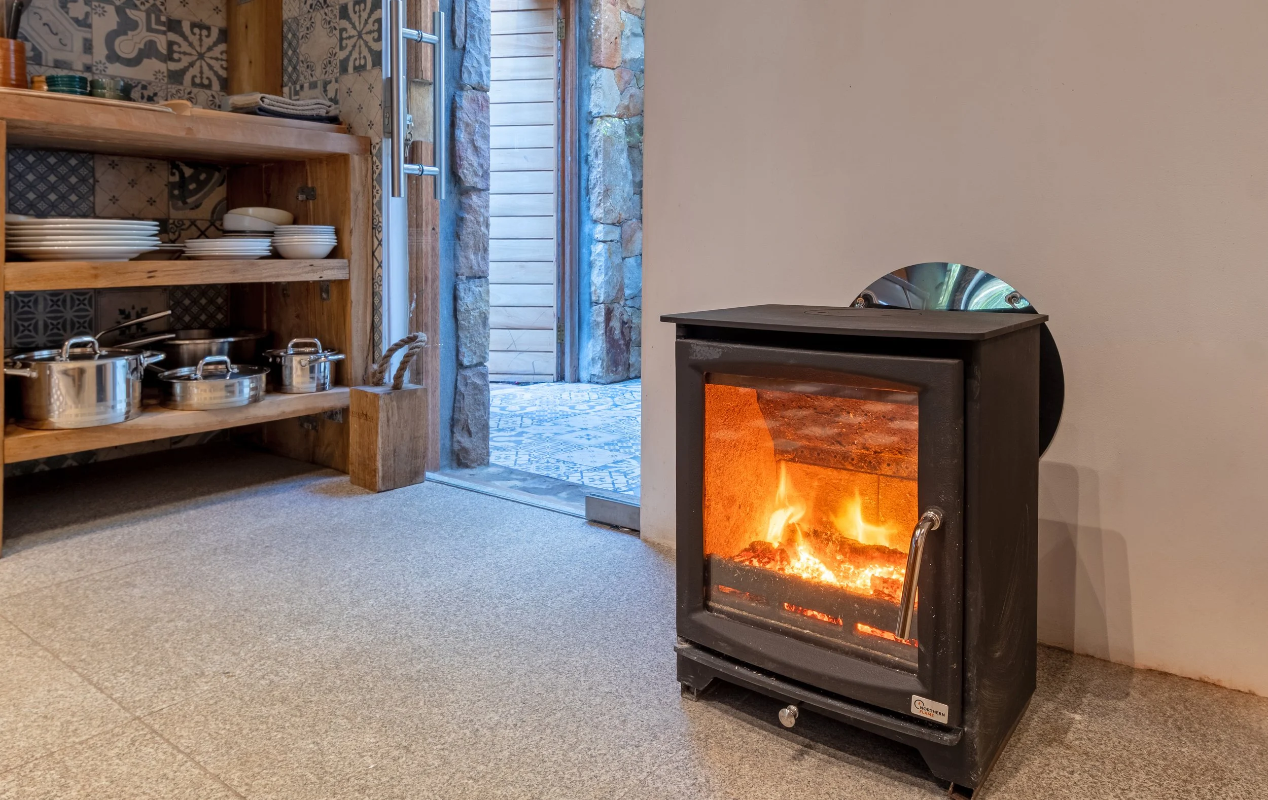 A cozy room with a wood-burning stove with a fire inside, a wooden shelf filled with plates and pots, and an open door leading outside to a patio area with stone and wood decor.
