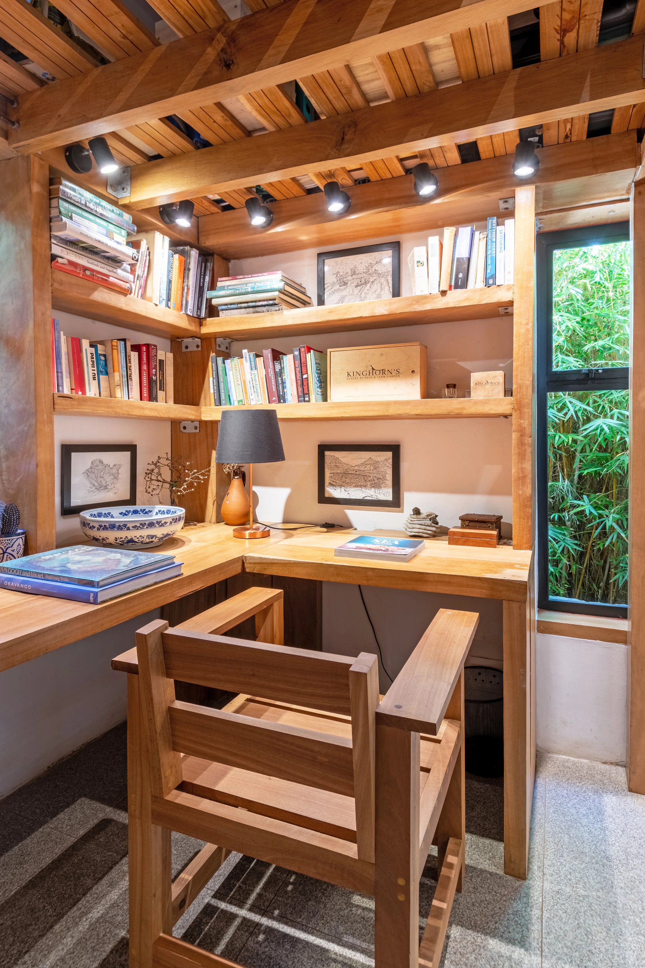 Small wooden study nook with built-in desk, surrounded by wooden shelves filled with books, a small table lamp, framed artwork, and a window with greenery outside.