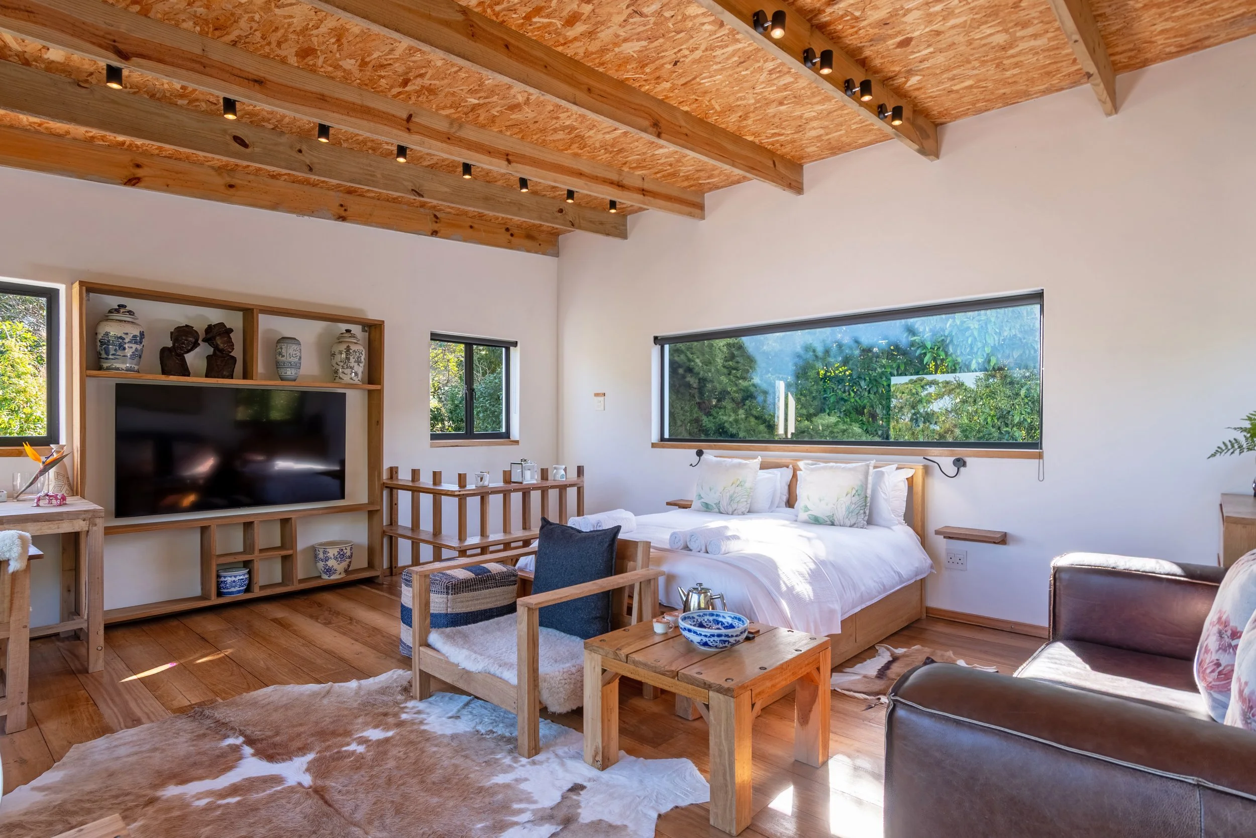 Cozy living room with white walls, wooden ceiling and floor, featuring a bed, chairs, a TV, shelves with pottery and art, and large windows with view of greenery outside