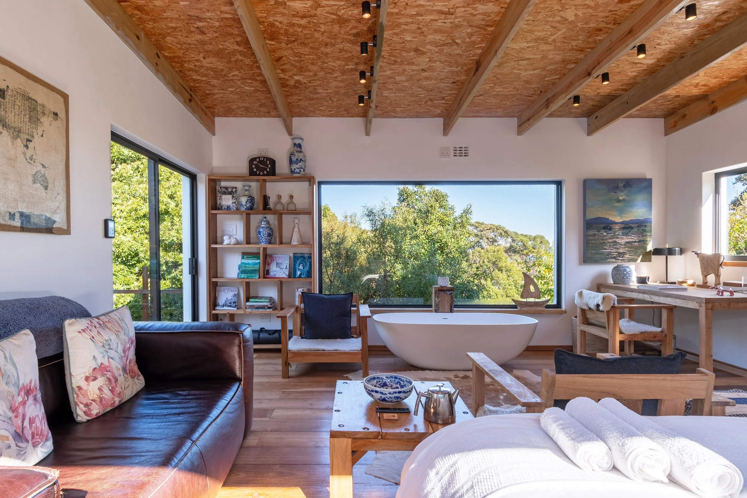 Interior of a modern, rustic-style bedroom with a large window overlooking trees, a free-standing bathtub, a leather sofa with floral pillows, a wooden side table with towels, a bookshelf, and decorative items and artwork.