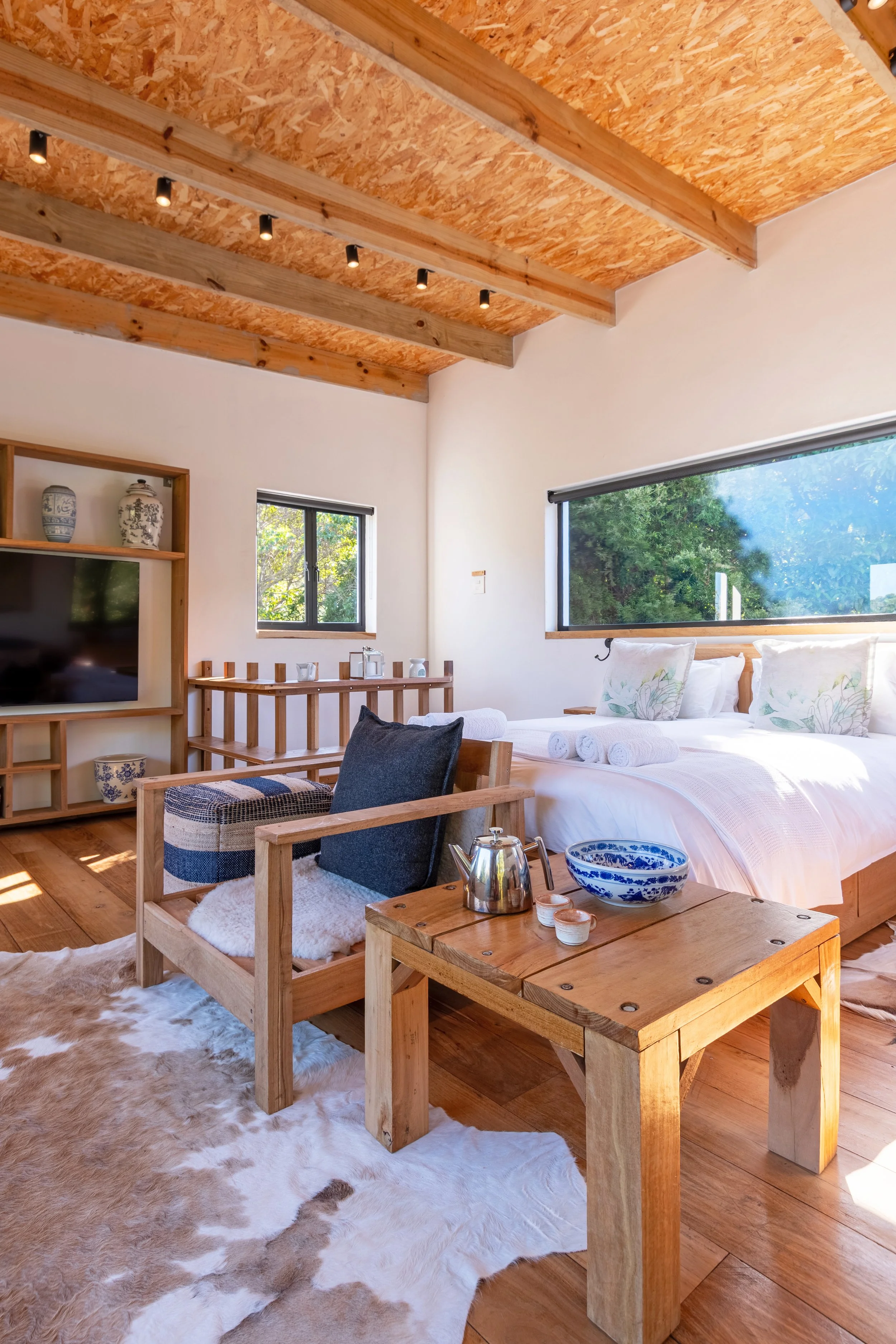 A rustic bedroom with a vaulted wooden ceiling and large windows, featuring a bed with rolled towels, a wooden table with a teapot and bowl, a cozy armchair, a cowhide rug, and a wooden shelf with decorative vases.
