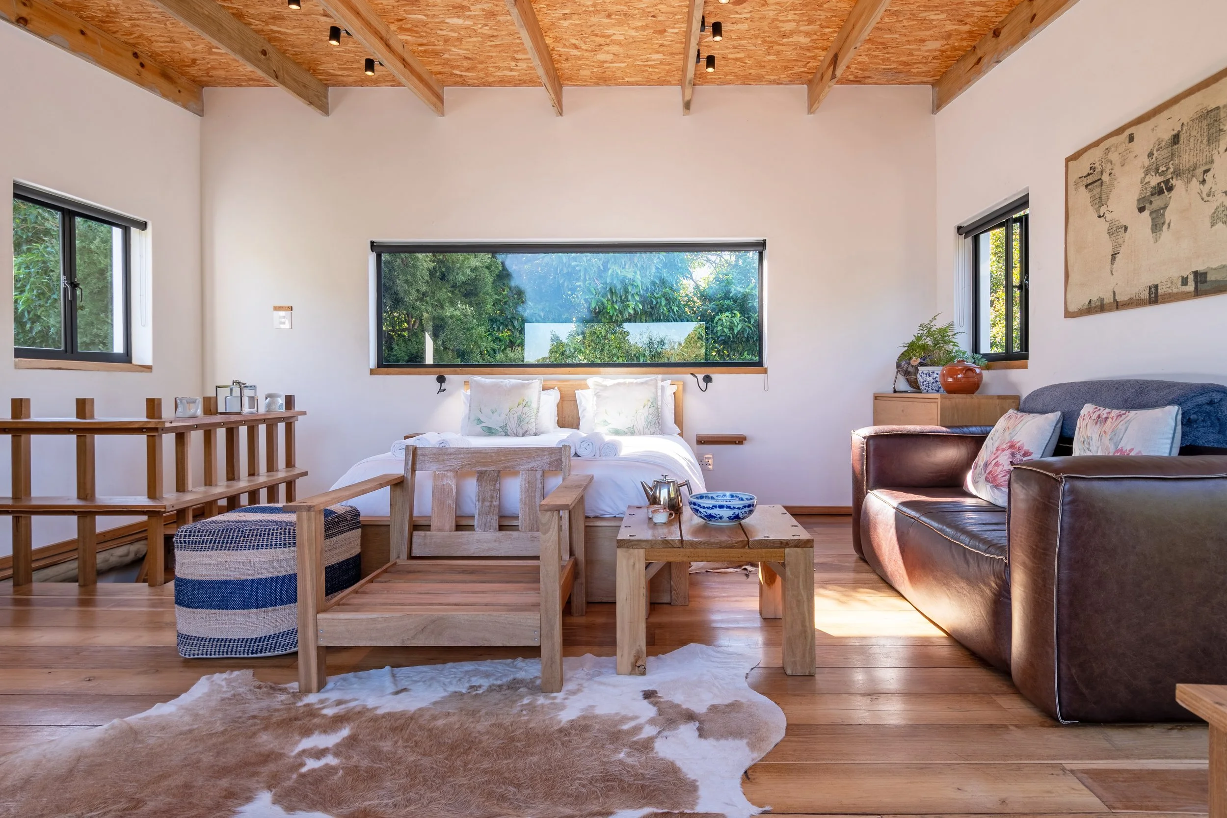 Bright and airy bedroom with a wooden ceiling, large windows, a bed with white bedding, a leather sofa, wooden furniture, a cowhide rug, and green plants.