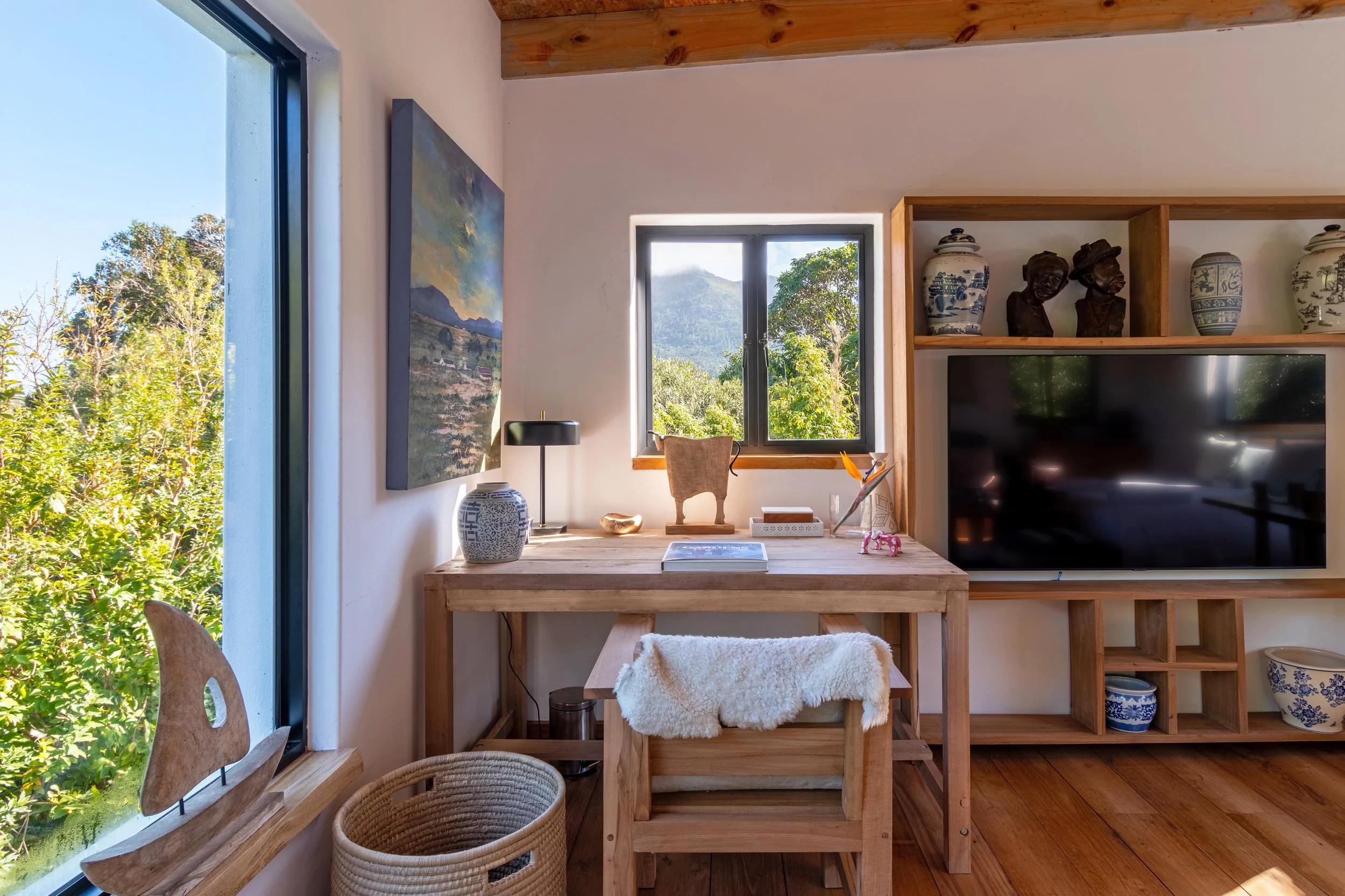 Cozy home office with wooden desk, window view of green trees and mountains, bookshelf with decorative vases and busts, and a large flat-screen TV.