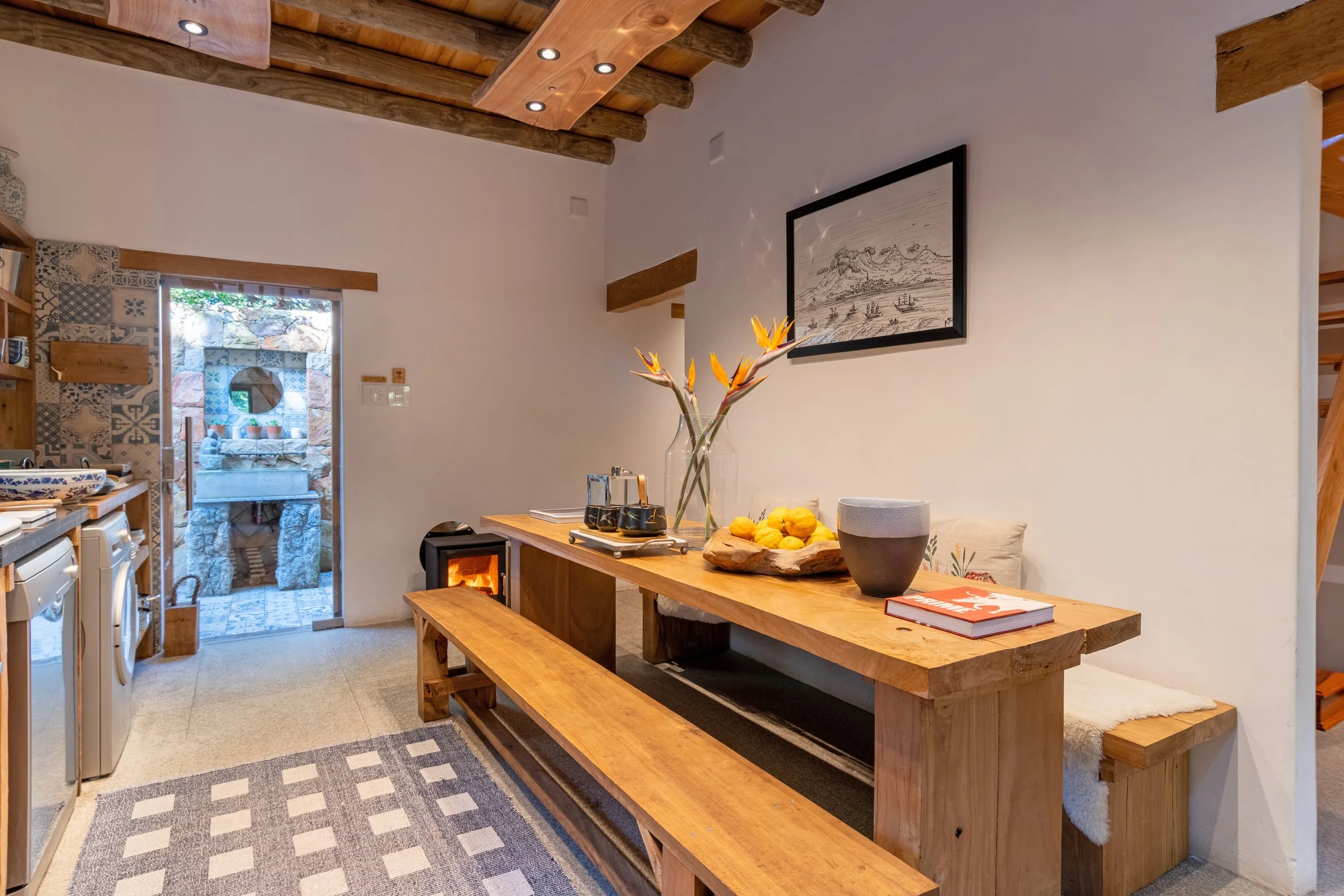 Dining area with a wooden table and bench, decorative items, and a wall art, next to a laundry area with washing machines, and an outdoor stone oven visible through the doorway.