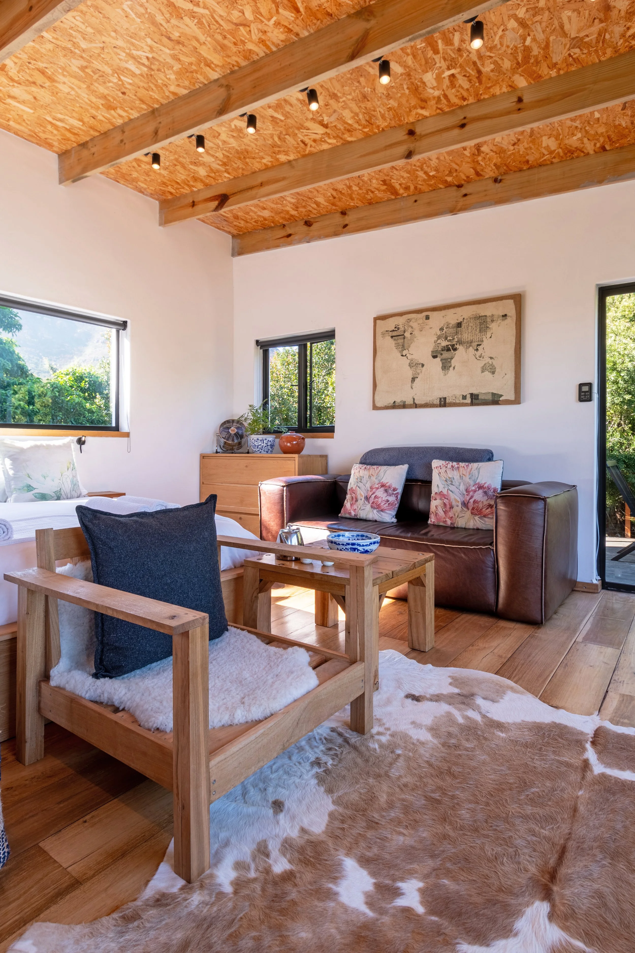 A living room with wooden floors, a cowhide rug, a wooden chair with a fur cushion, a brown leather sofa with floral pillows, and a light wood sideboard near two windows with black frames, decorated with plants and pottery, and a wall map.