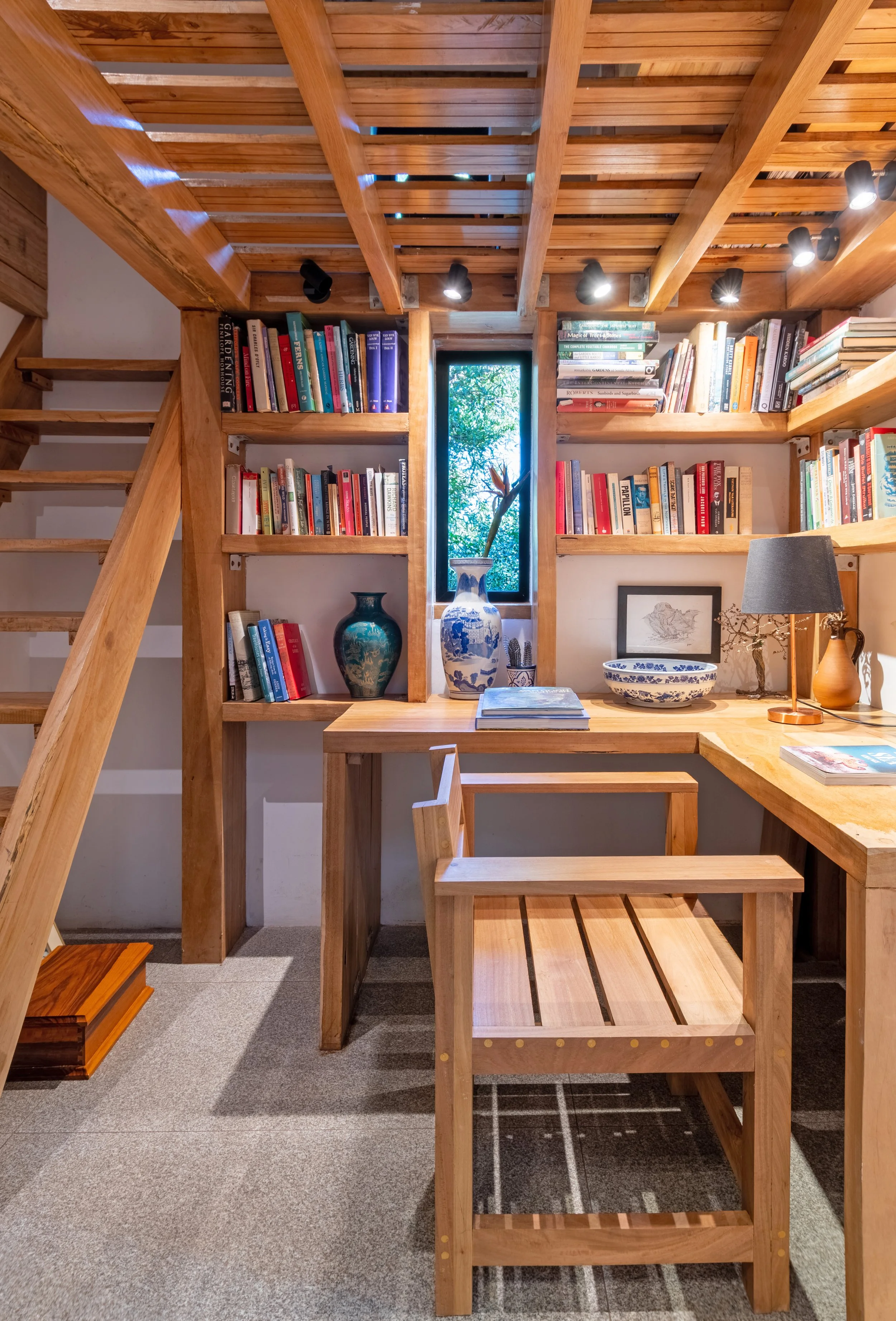 A cozy wooden study corner with built-in shelves filled with books, a small desk, a chair, decorative vases, a framed sketch, a bowl, and a table lamp, illuminated by track lighting and a small window showing green foliage outside.