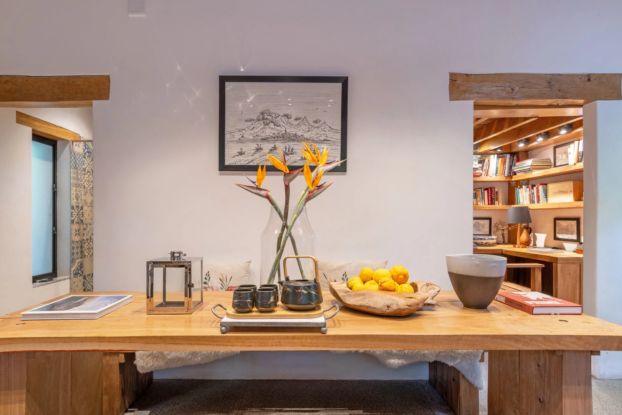 A wooden table decorated with a vase of Bird of Paradise flowers, a wooden bowl of yellow lemons, and various cups and decor items, set against a white wall with framed art and a bookshelf in the background.