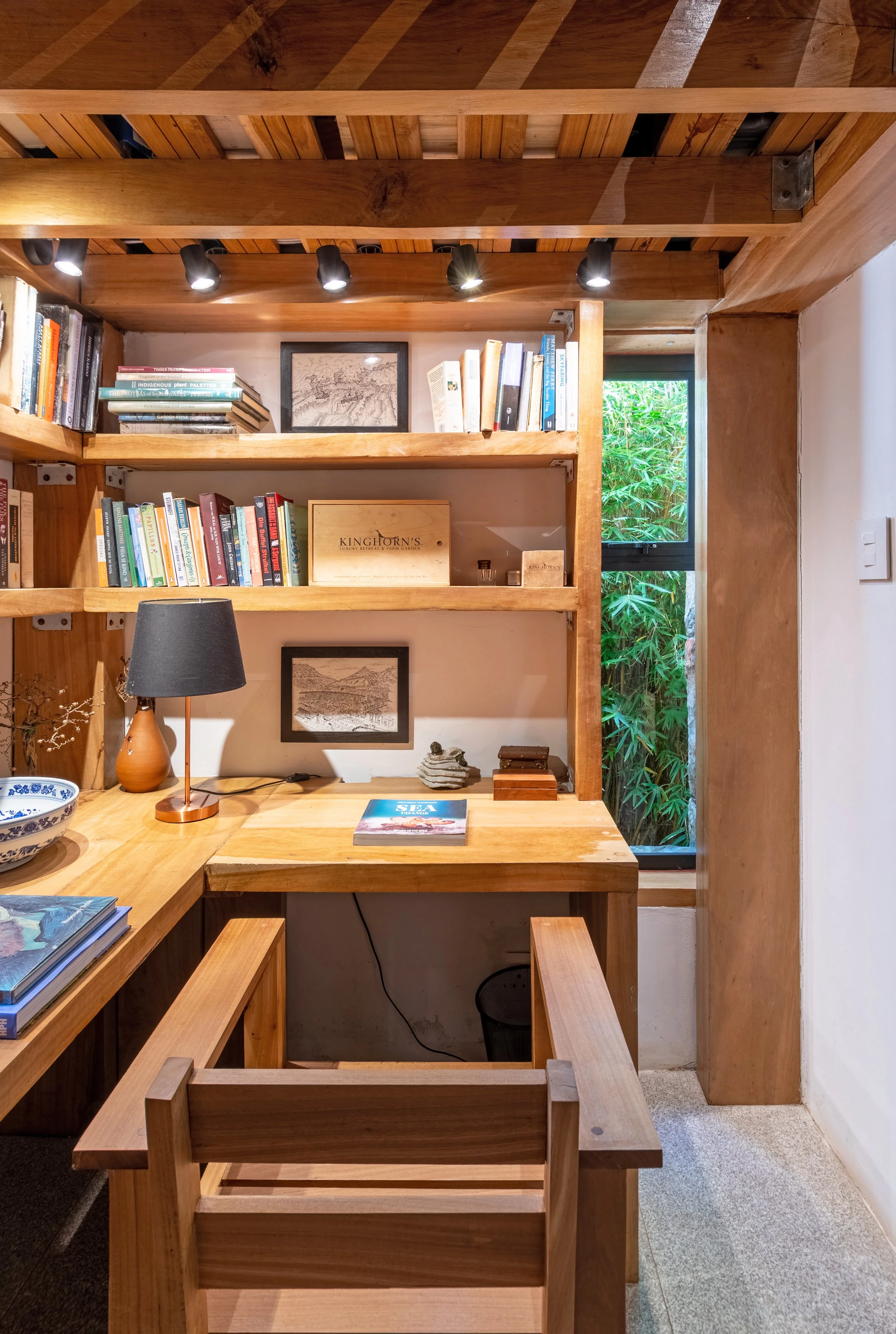 A cozy wooden home office with built-in bookshelves, a desk, a black lamp, and a view of outside greenery through a window.