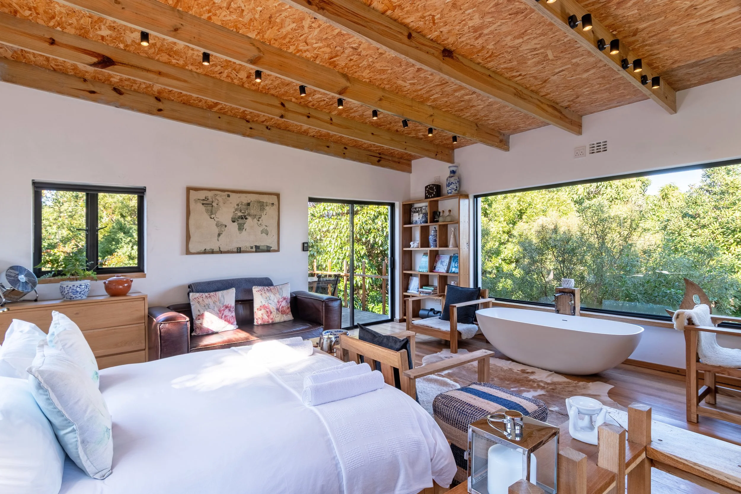 Bright bedroom with large window and greenery view, featuring a white bed, a wooden drawer with decorative items, a leather sofa with floral pillows, a wooden shelf with decorative objects, a freestanding bathtub near the window, and wooden ceiling b