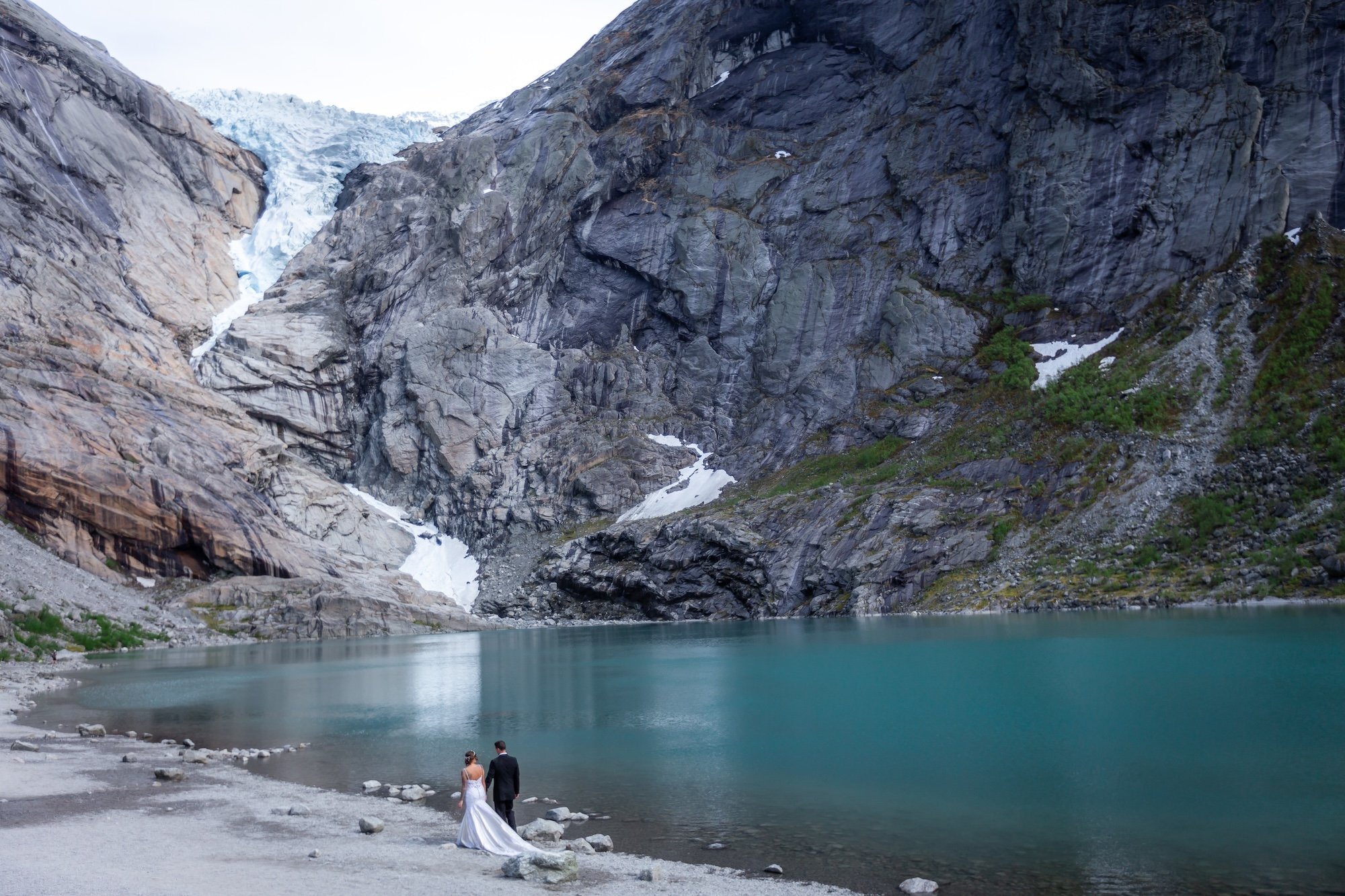 A bride and groom standing by a turquoise lake surrounded by steep rocky cliffs and patches of green vegetation.