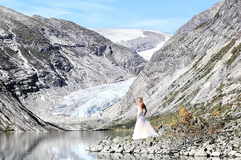 A woman in a white dress standing on rocks by a lake in a mountainous area with a glacier and snow-capped peaks in the background.