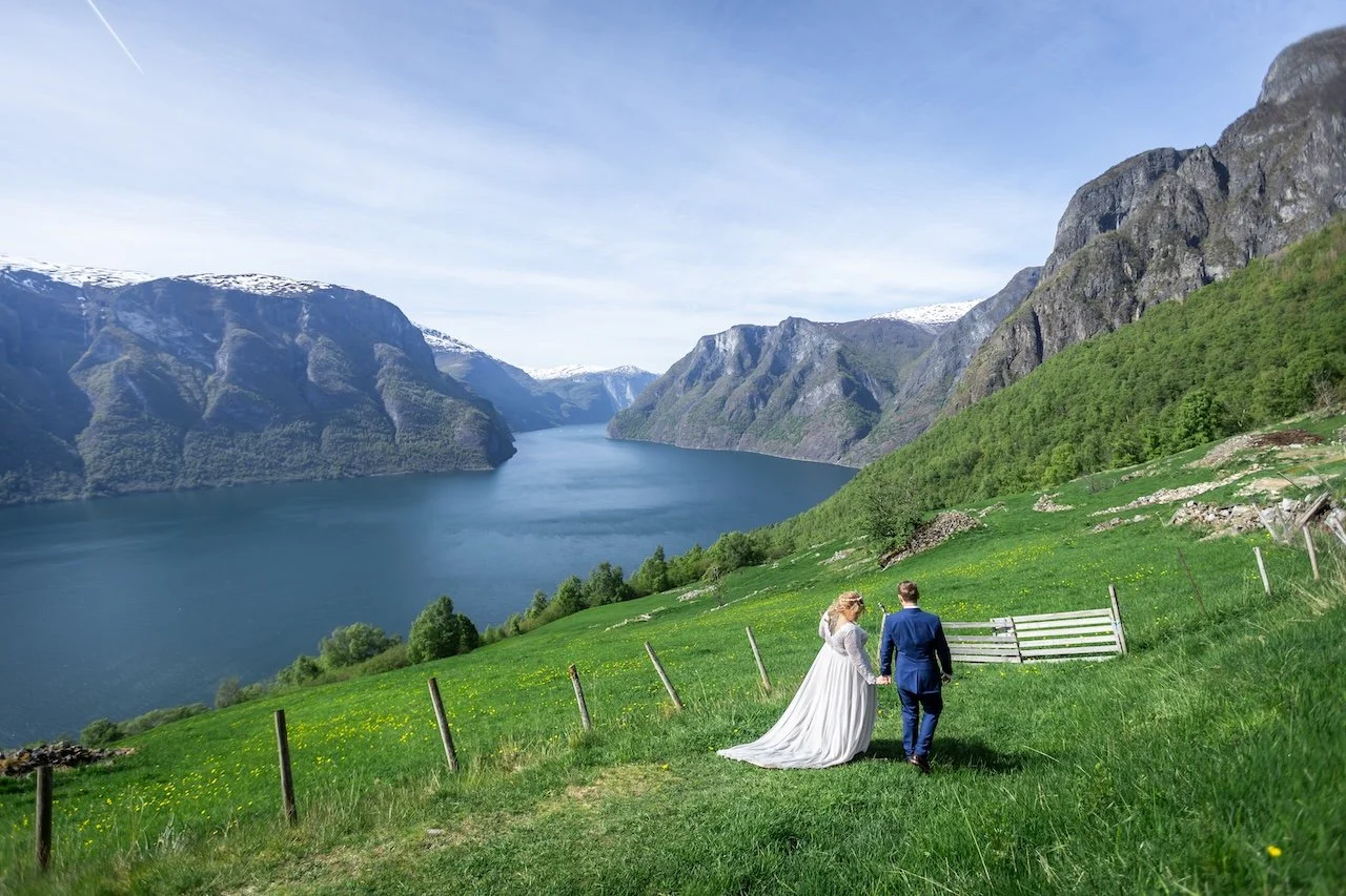 A bride and groom walking hand in hand on a green grassy hillside overlooking a fjord with steep mountains and snow-capped peaks in the background.