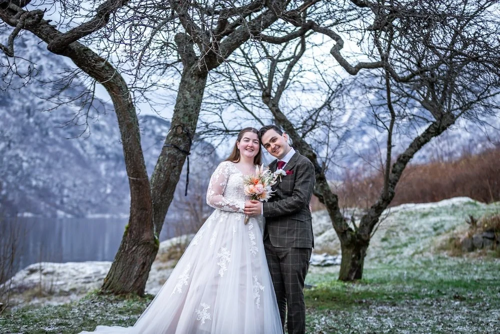 A bride and groom smiling and standing closely together outdoors in a winter landscape with snow, leafless trees, and mountains in the background. The bride is holding a bouquet of flowers and wearing a white wedding dress, while the groom is in a gr