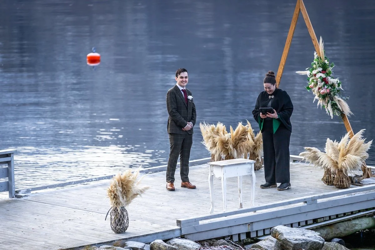 A couple getting married on a dock by a lake with a wooden arch decorated with flowers and pampas grass, a white table, and a vase of pampas grass in the foreground.