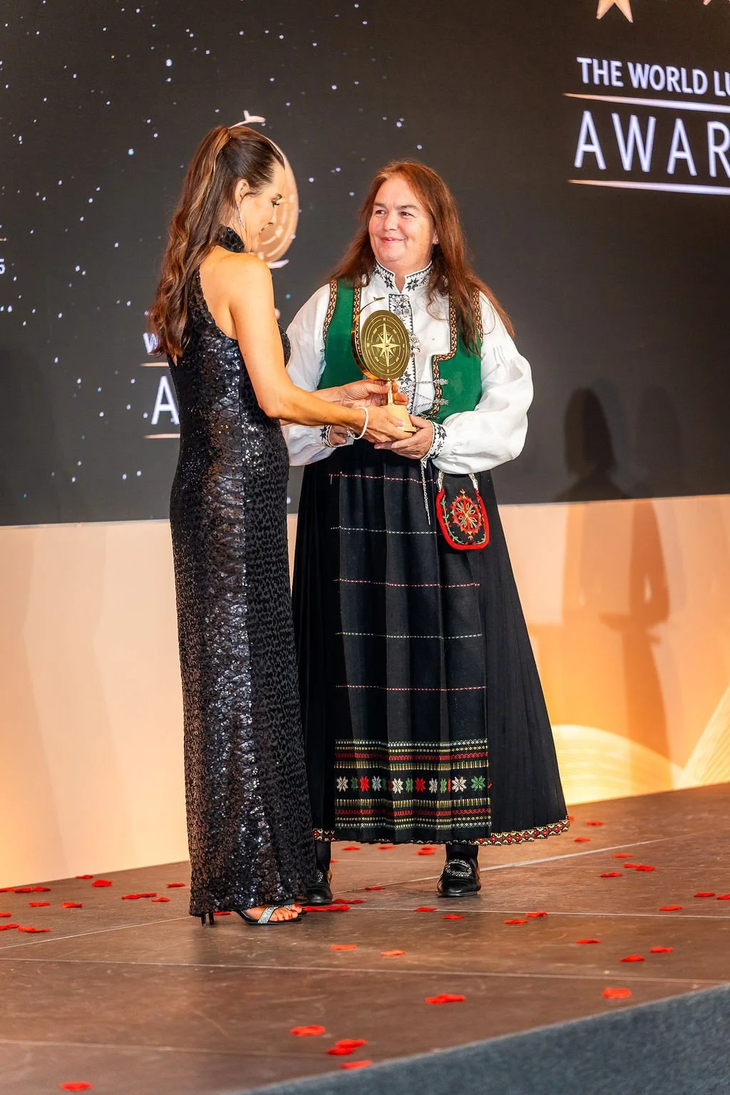A woman in a black sequin dress receives an award from another woman in traditional clothing on stage at an award ceremony, with a backdrop that says 'The World Leadership Awards'.