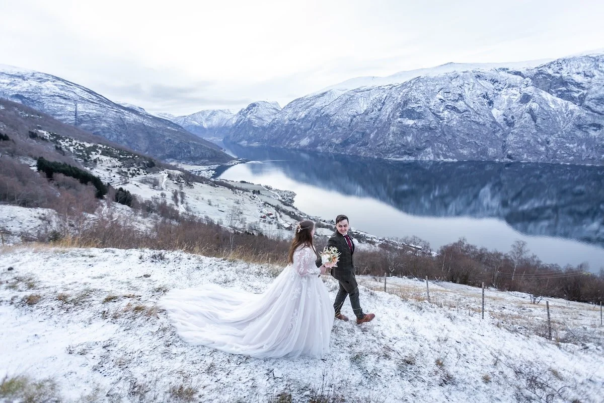 A bride and groom walking outside in a snowy landscape with mountains and a lake in the background.
