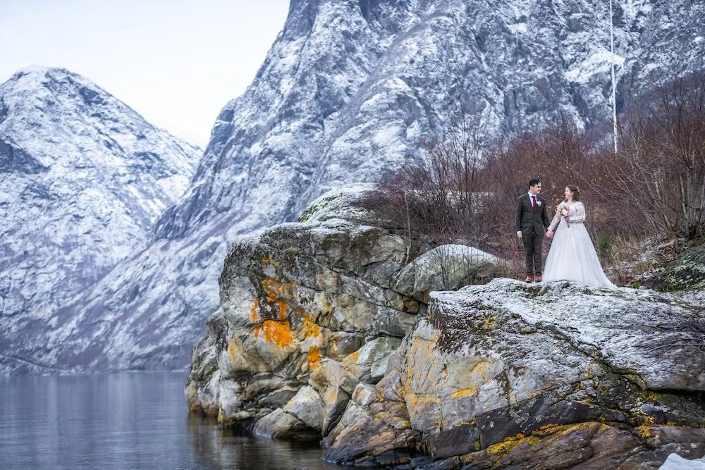 A bride and groom holding hands standing on a large rocky outcrop near a body of water with snow-covered mountains in the background.