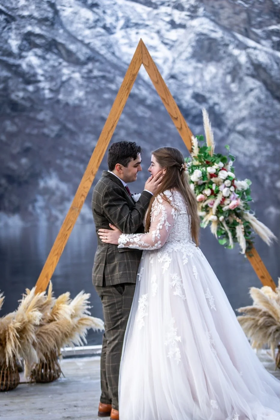 A bride and groom holding hands and touching faces during their wedding in front of a wooden triangular arch decorated with flowers, with a mountain and lake in the background.