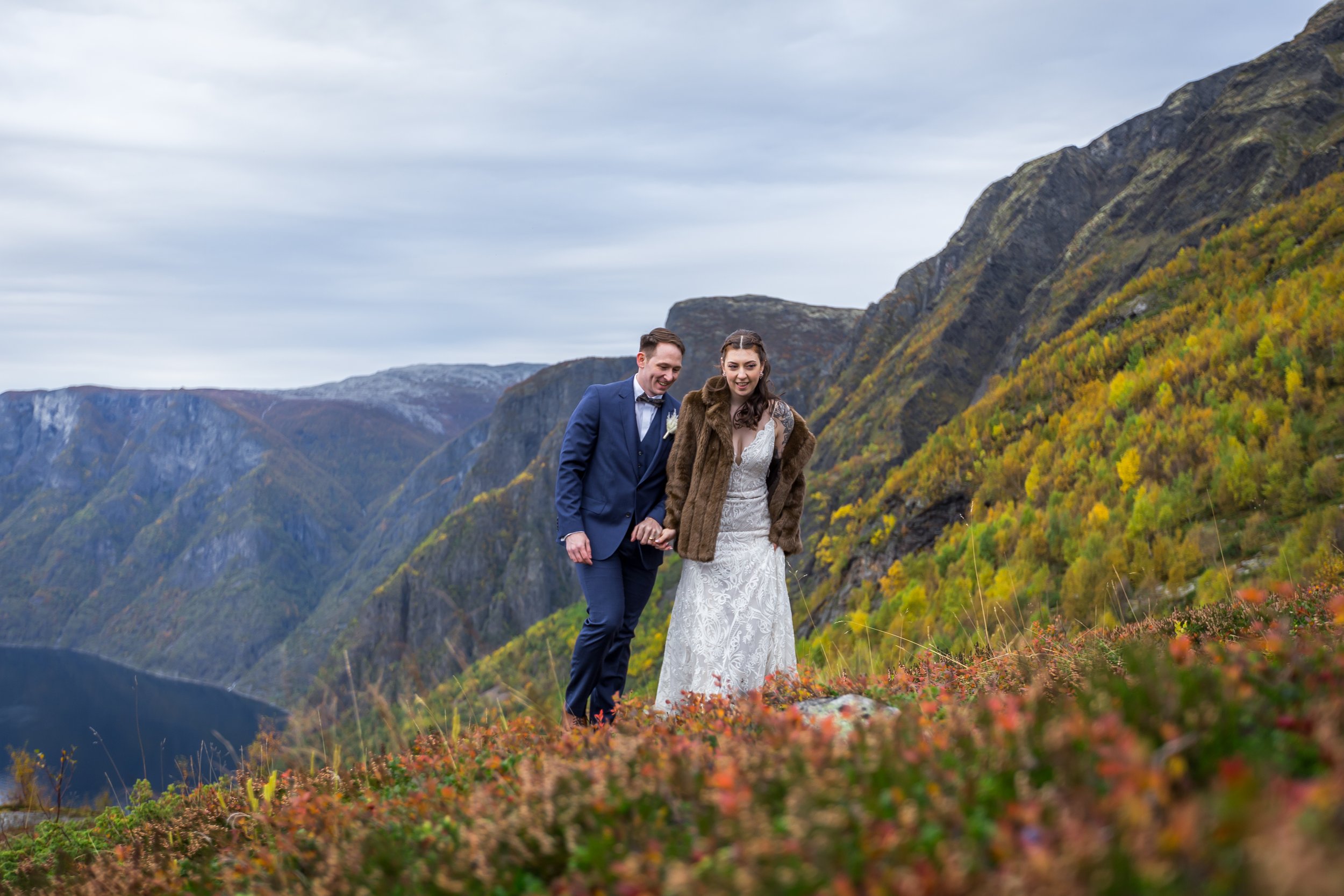 A couple dressed in wedding attire walking outdoors in front of scenic mountains with colorful autumn foliage.