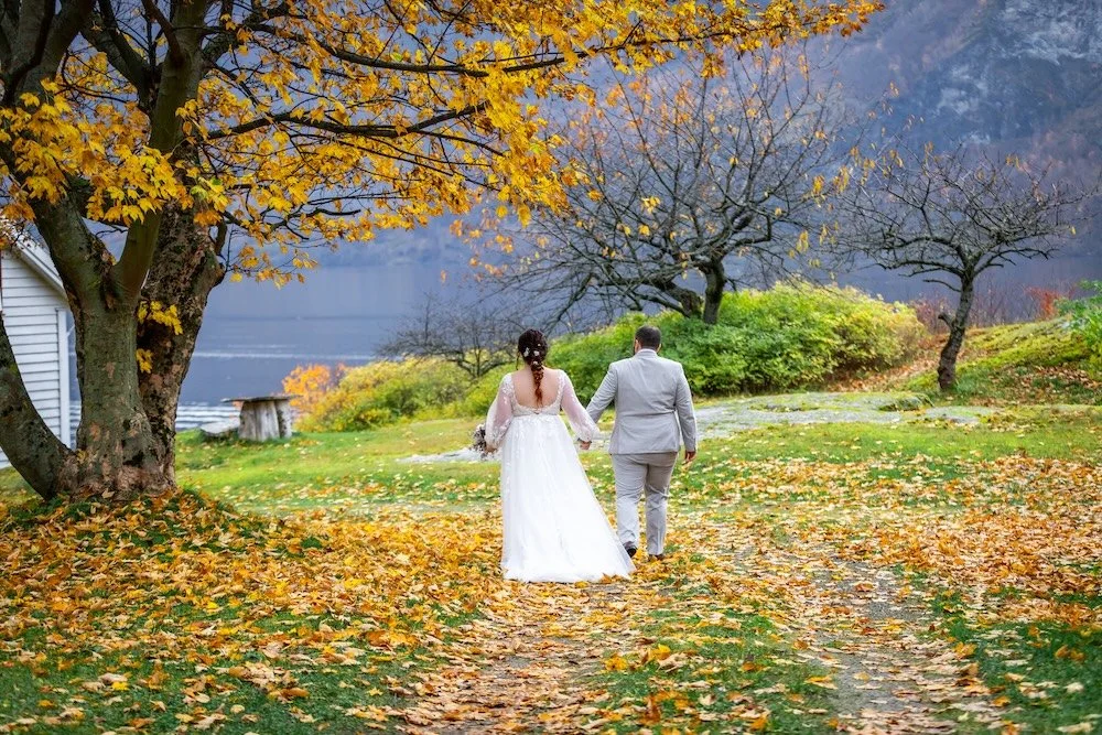 A bride and groom walking hand in hand on a leaf-covered path in an outdoor setting with trees, some in fall foliage, and a mountain in the background.