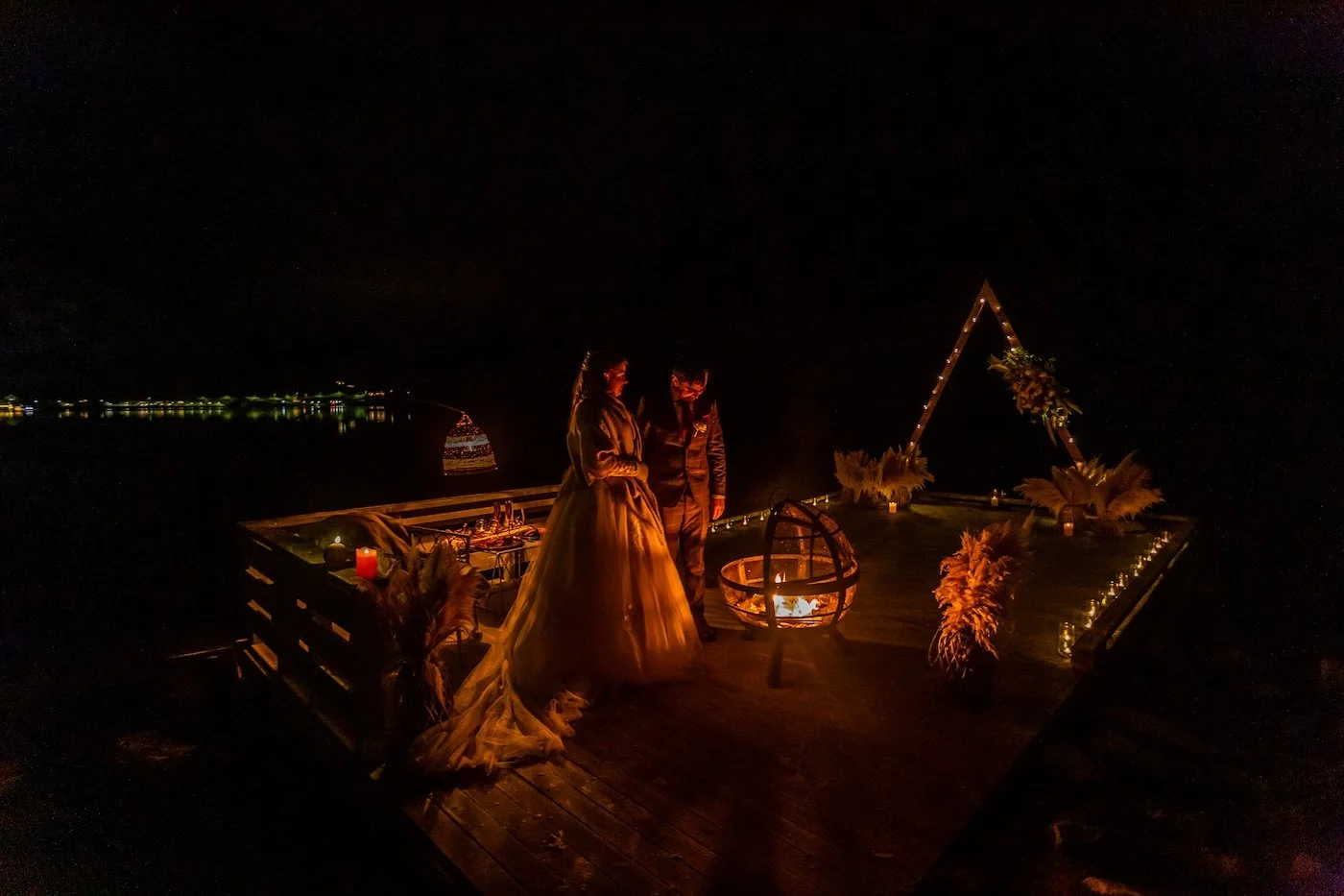 A couple stands on a wooden pier at night, surrounded by candles and floral decorations, with an open fire and a lit triangular wedding arch in the background over a body of water.