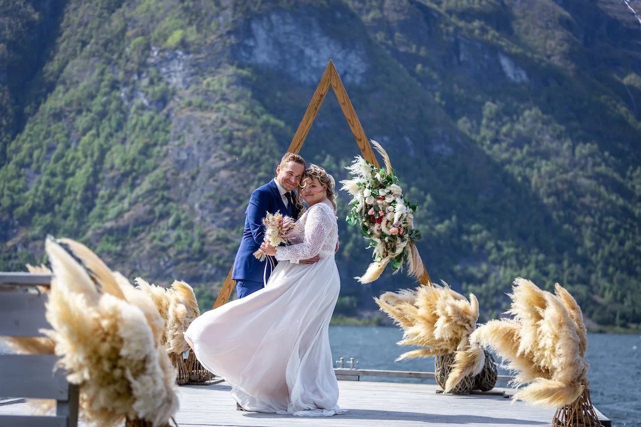 A couple in wedding attire embracing outdoors on a wooden dock with a mountain lake background, decorated with floral arrangements and pampas grass.