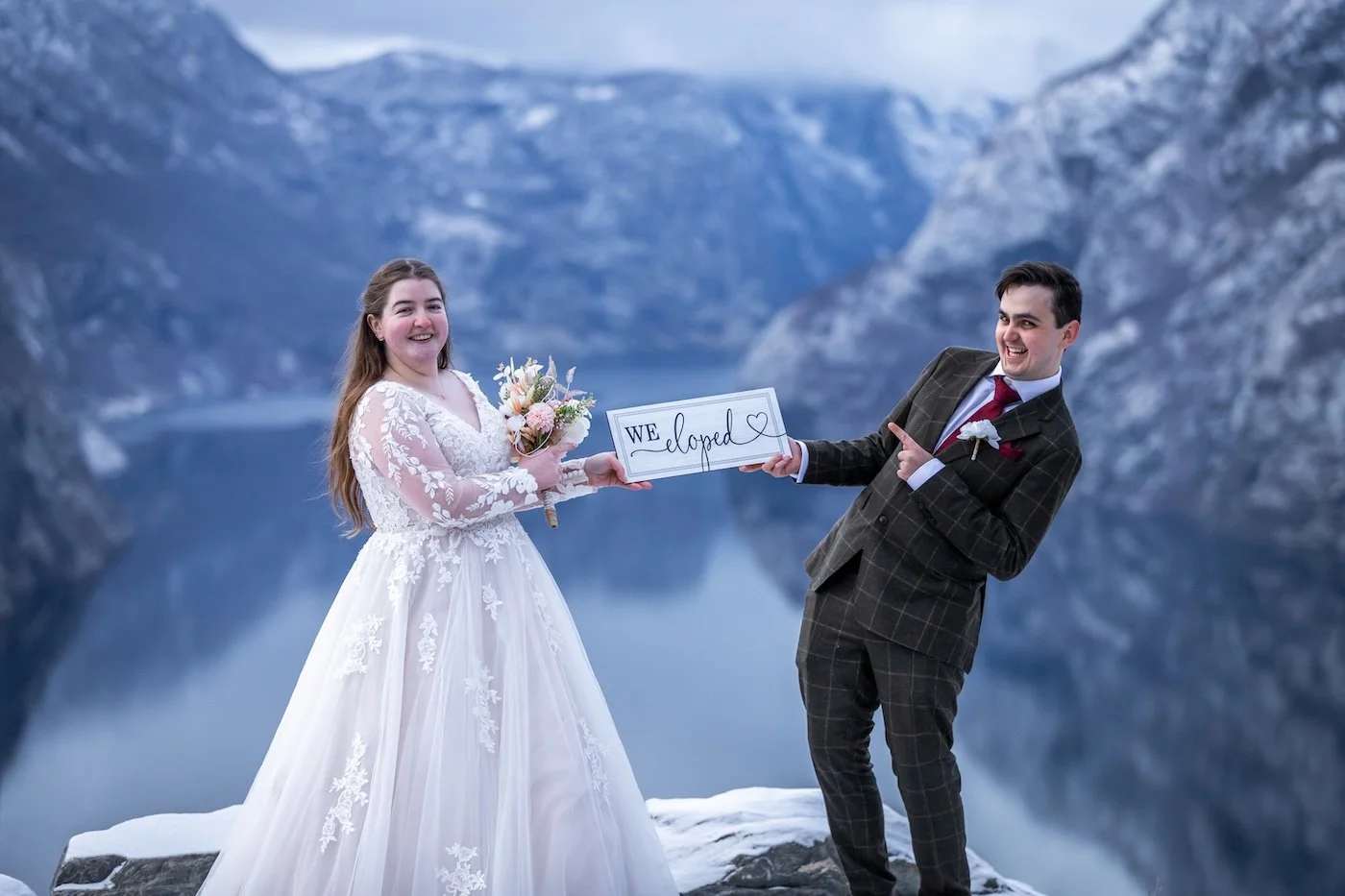A newlywed couple stands outdoors by a body of water with mountains in the background. The bride is wearing a white wedding dress with lace details and holding a bouquet of flowers. The groom is dressed in a brown checkered suit with a white shirt an