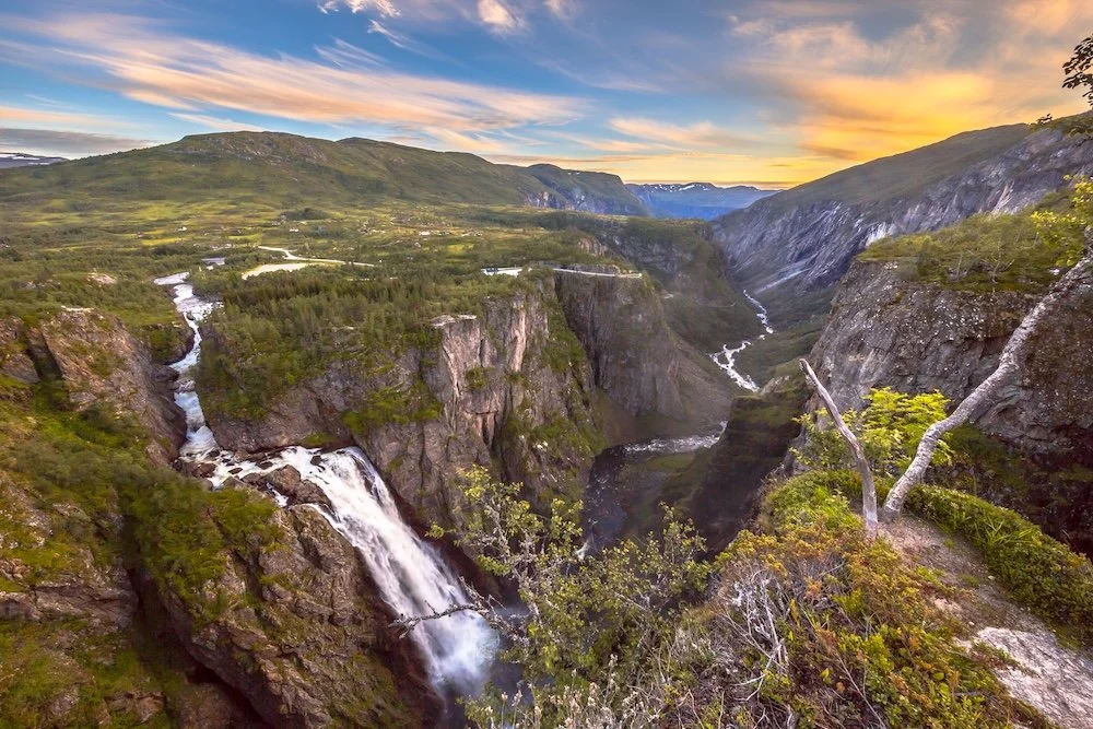 A scenic view of a canyon with waterfalls, green trees, and a river, under a colorful sky at sunset.
