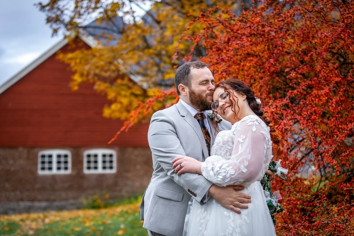 A happy couple in wedding attire embracing outdoors during autumn with red and orange trees and a rustic barn in the background.