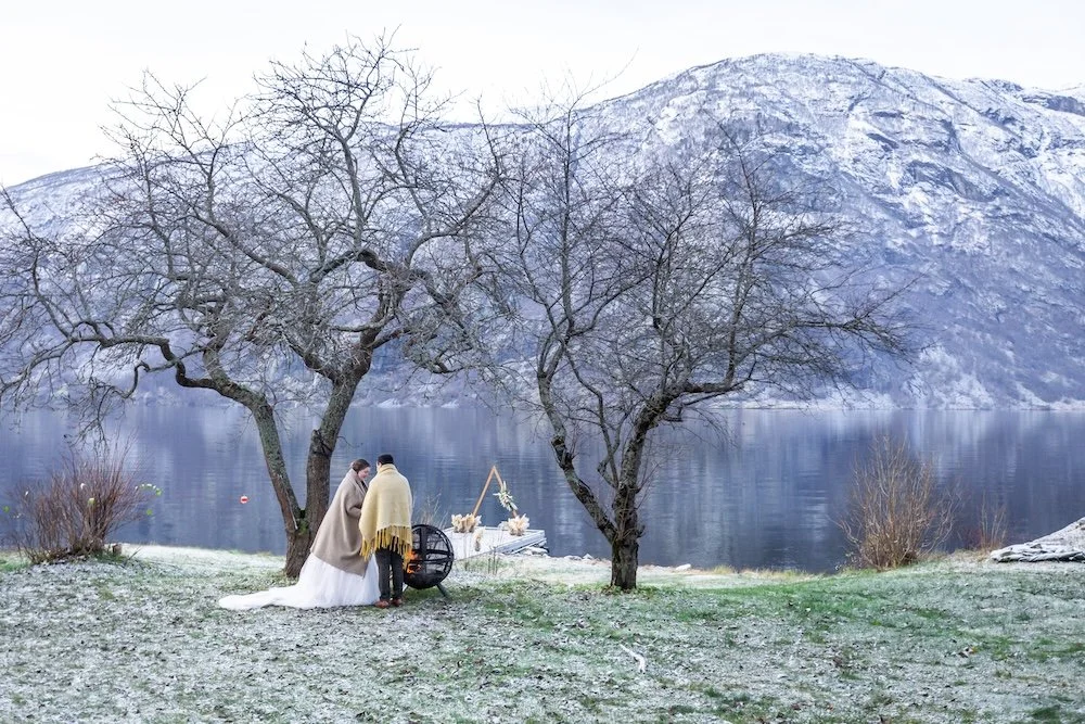 A couple gets married outdoors by a lake with snow-capped mountains in the background, under leafless trees, with a small decorated altar nearby.