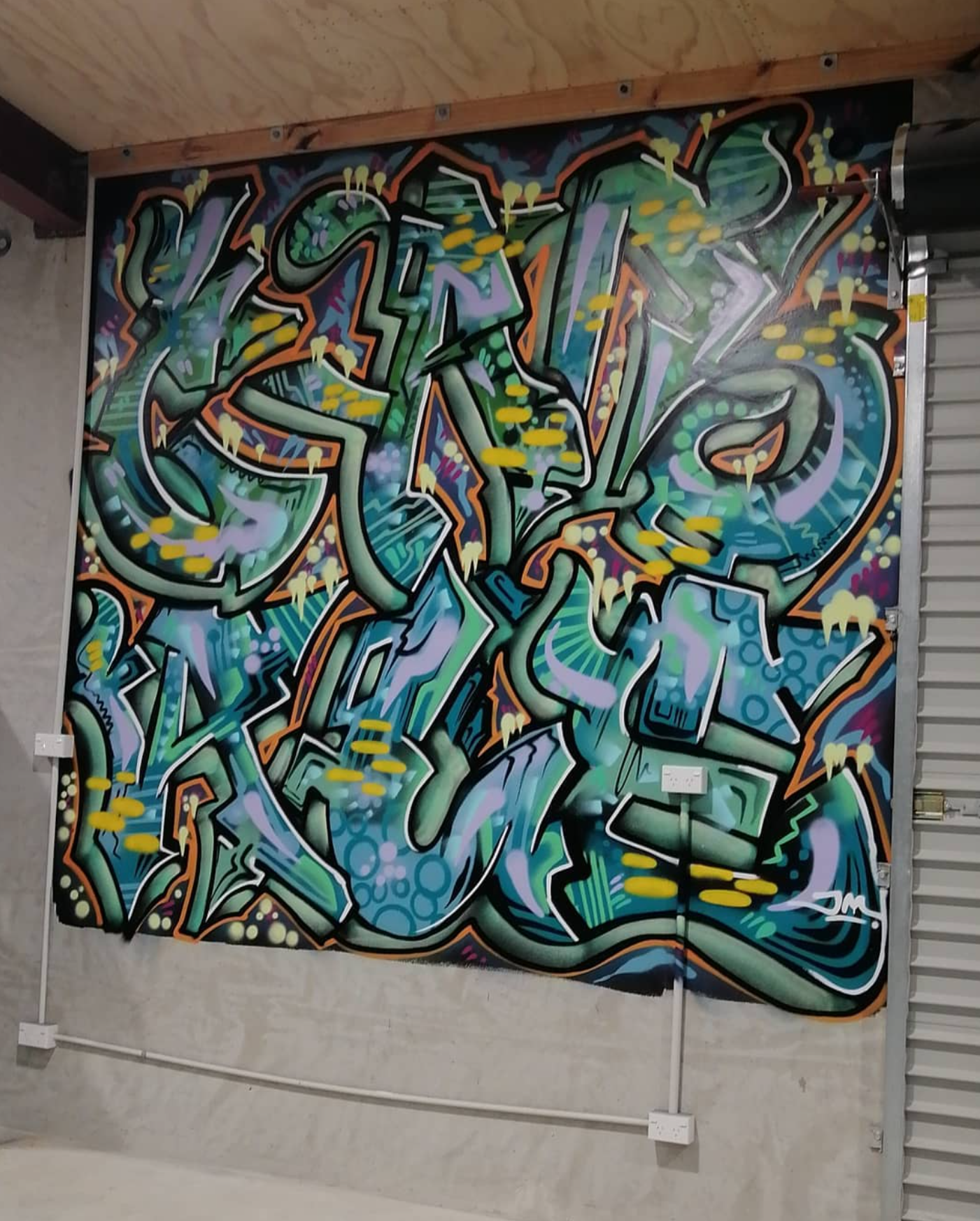 Colorful graffiti mural on a wall with vibrant abstract designs, shapes, and patterns, including arrows, circles, and lines, with a small signature in the bottom right corner.