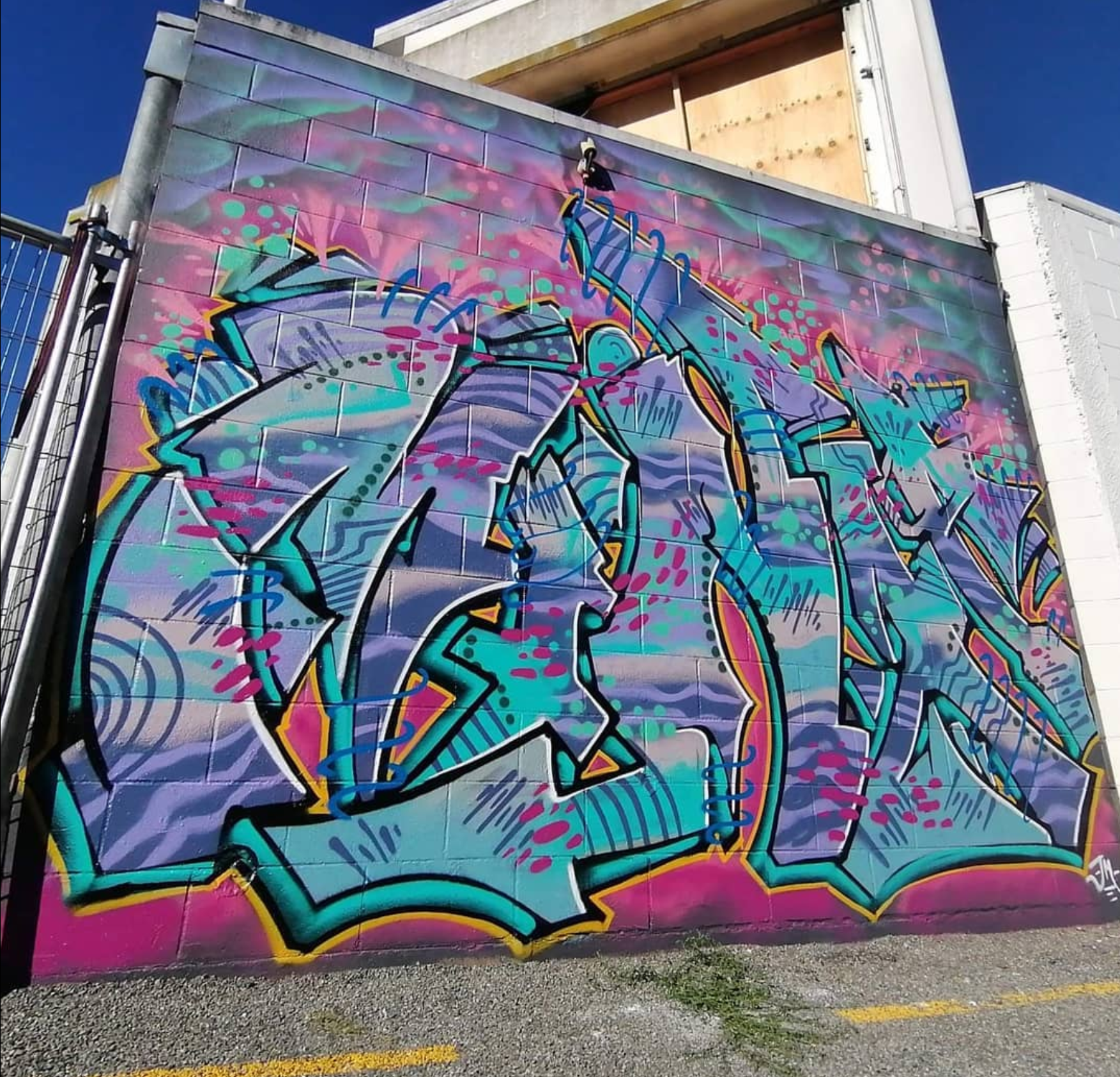 Colorful graffiti mural on a brick wall featuring abstract shapes and vibrant colors including pink, purple, blue, and green, with a clear blue sky overhead.