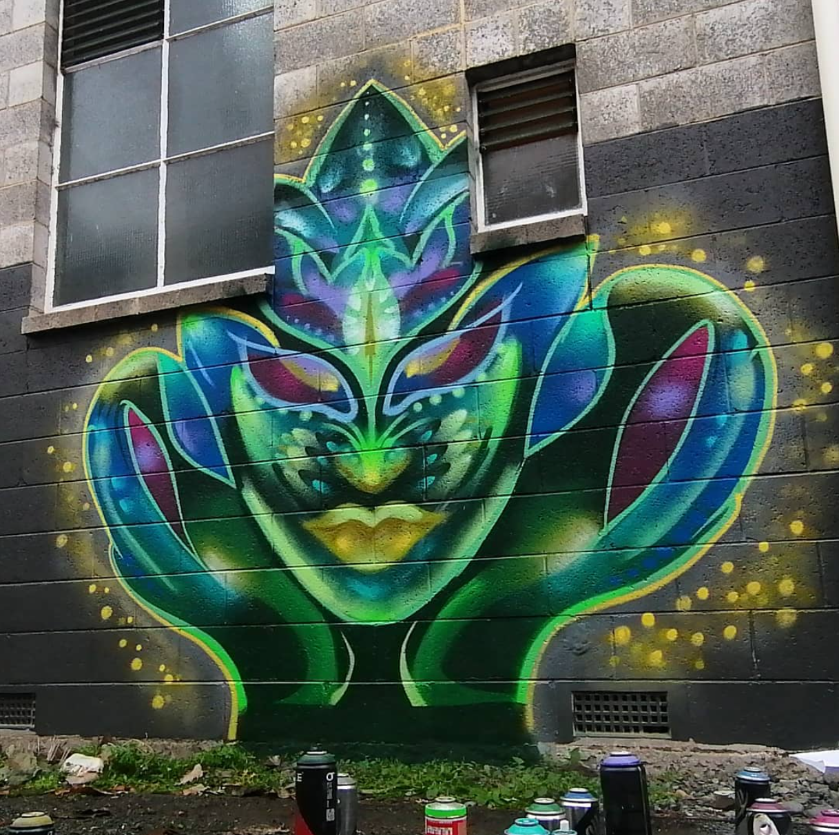 Colorful graffiti of a stylized face with feline features, green, blue, purple, and yellow accents, painted on a dark building wall near two windows.