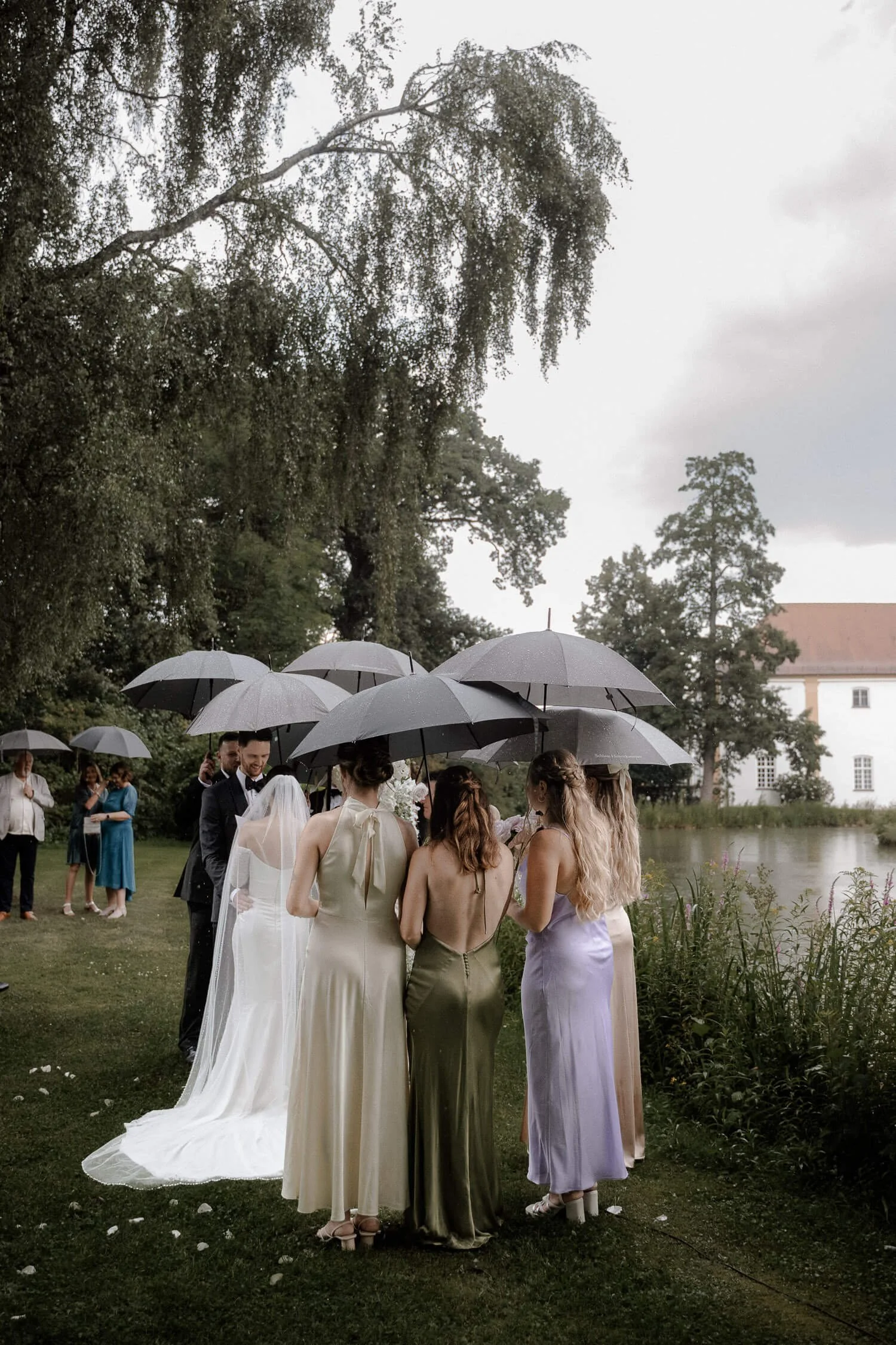 Hochzeitsfotograf München – freie Trauung am See mit Brautpaar und Gästen unter Regenschirmen