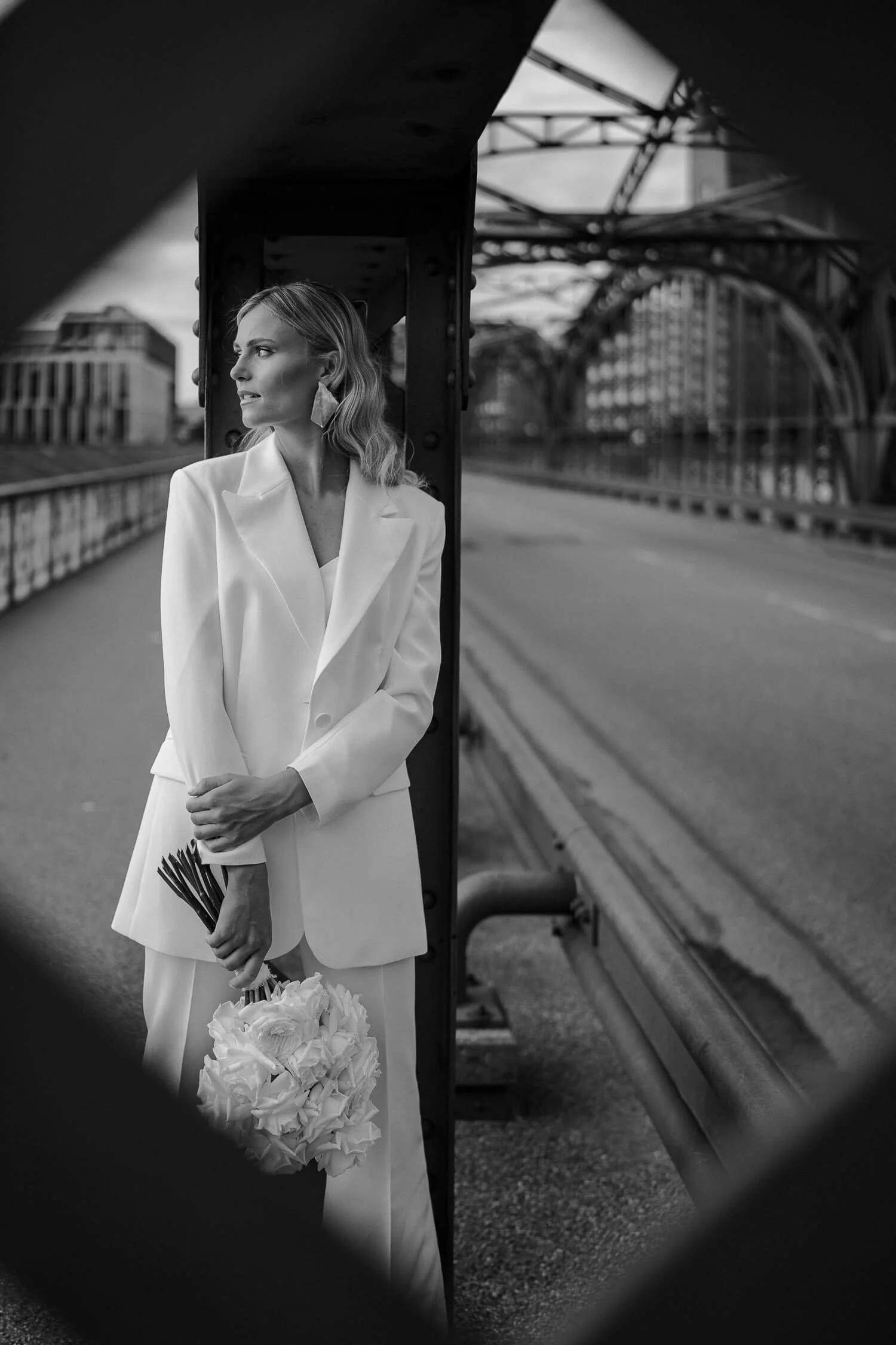 Moderne Braut in München – Stilvolles Hochzeitsfoto einer Frau im weißen Anzug auf urbaner Brücke.