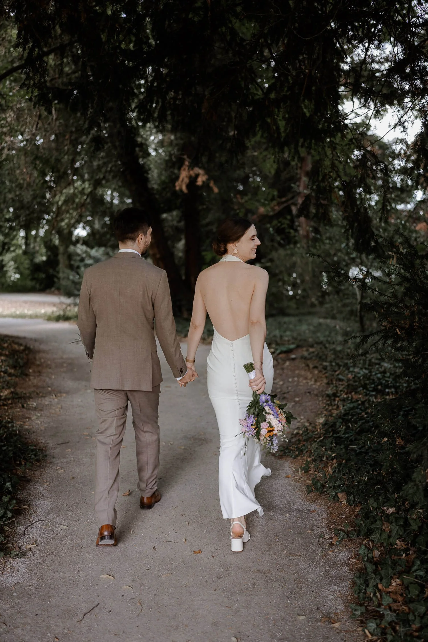 Brautpaar von hinten, Hand in Hand auf Waldweg, Braut schaut lächelnd zurück, Bouquet in der Hand – roseninsel hochzeit paarshooting spaziergang waldweg candid