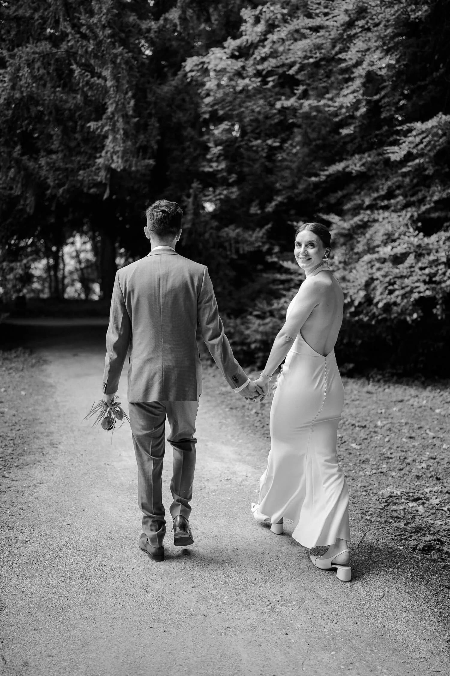 Schwarzweiß: Brautpaar von hinten, Hand in Hand auf Waldweg (Braut schaut zurück) – roseninsel hochzeit starnberger see spaziergang